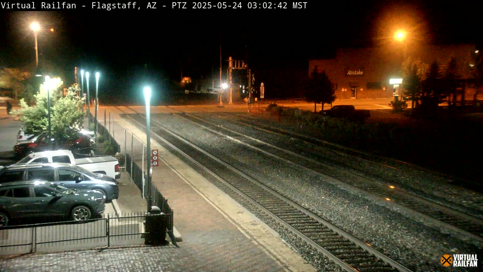 A nighttime view of train tracks at a station with several parked cars, streetlights, and a building in the background.