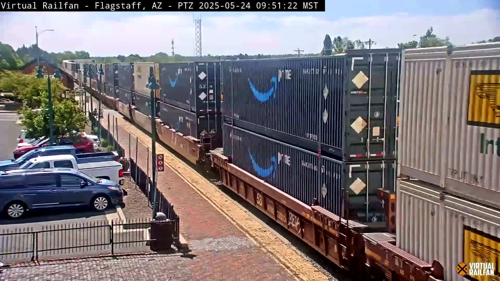 A long freight train carrying numerous shipping containers, some bearing the Amazon Prime logo, moves along a track beside a paved area with parked cars under a sunny sky.