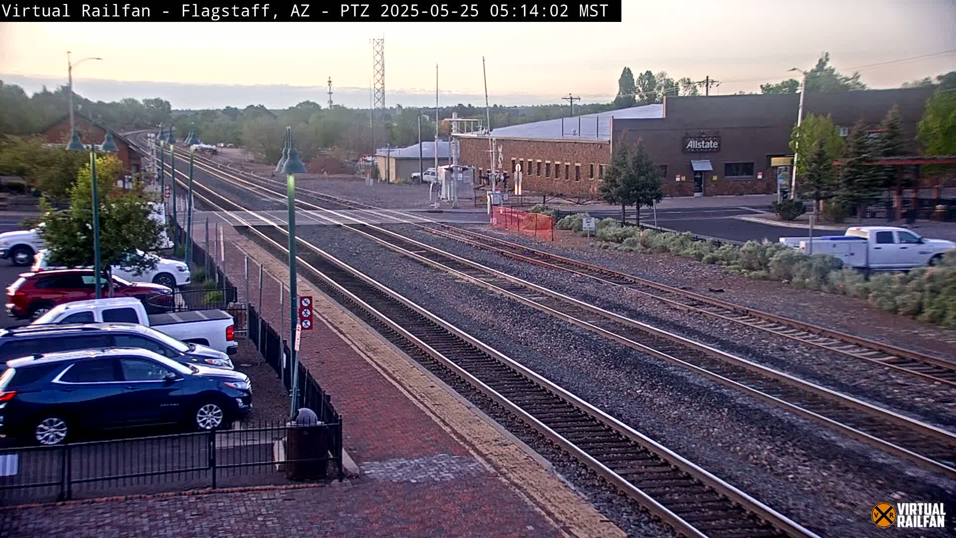 Multiple train tracks stretch into the distance past a parking lot with several cars, under a clear, dawn sky.