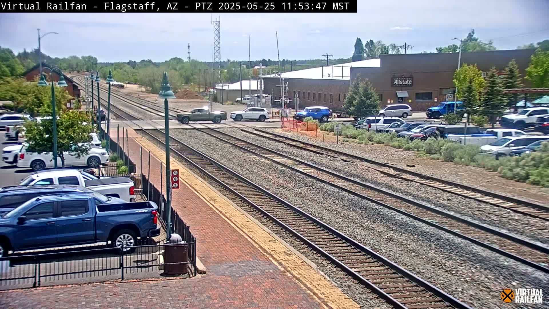 A sunny day shows multiple train tracks crossing a road where several vehicles are stopped and parked nearby buildings.