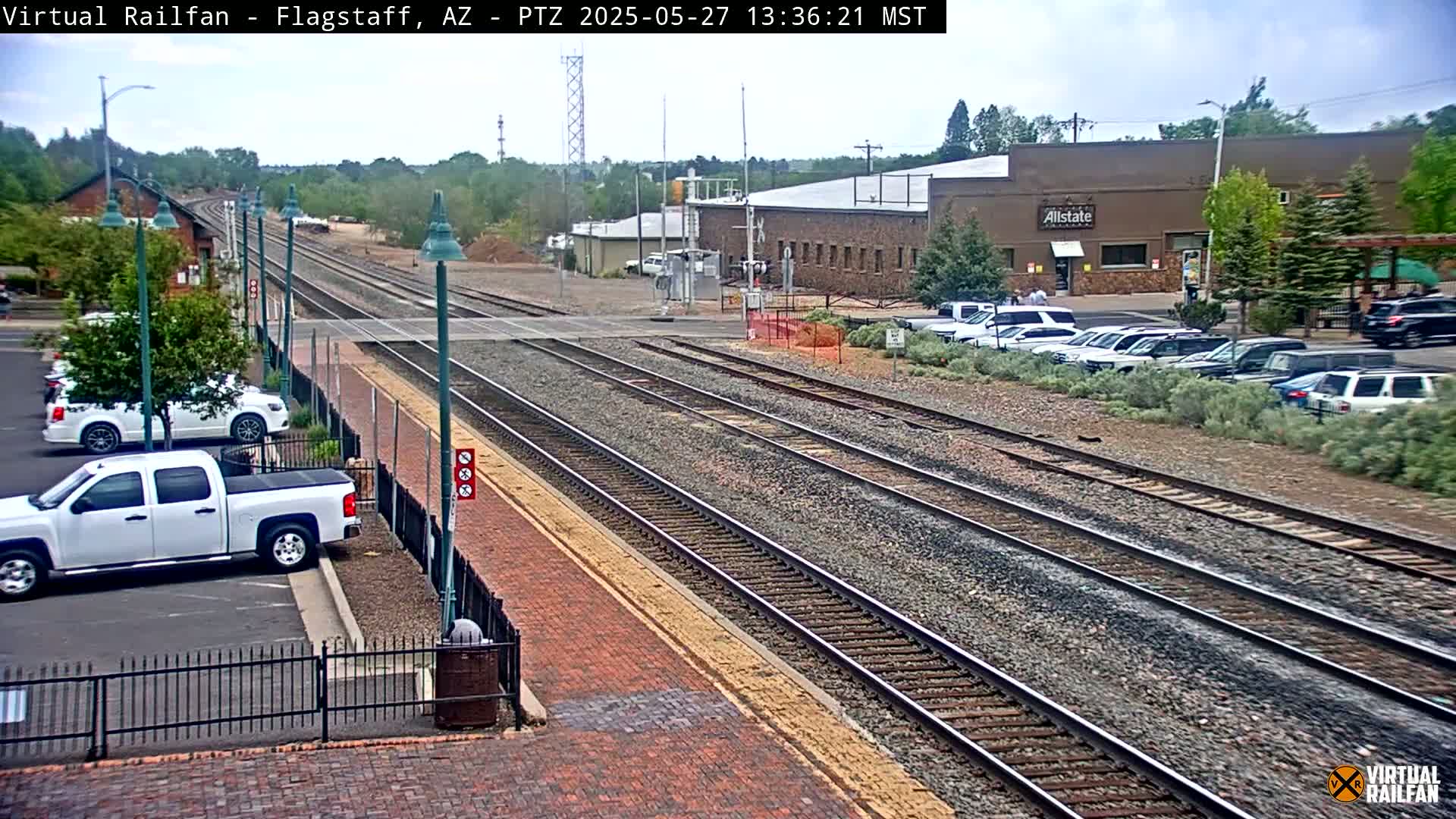 A mostly sunny day at a train station with multiple train tracks, several parked cars, and a brick building in the background.