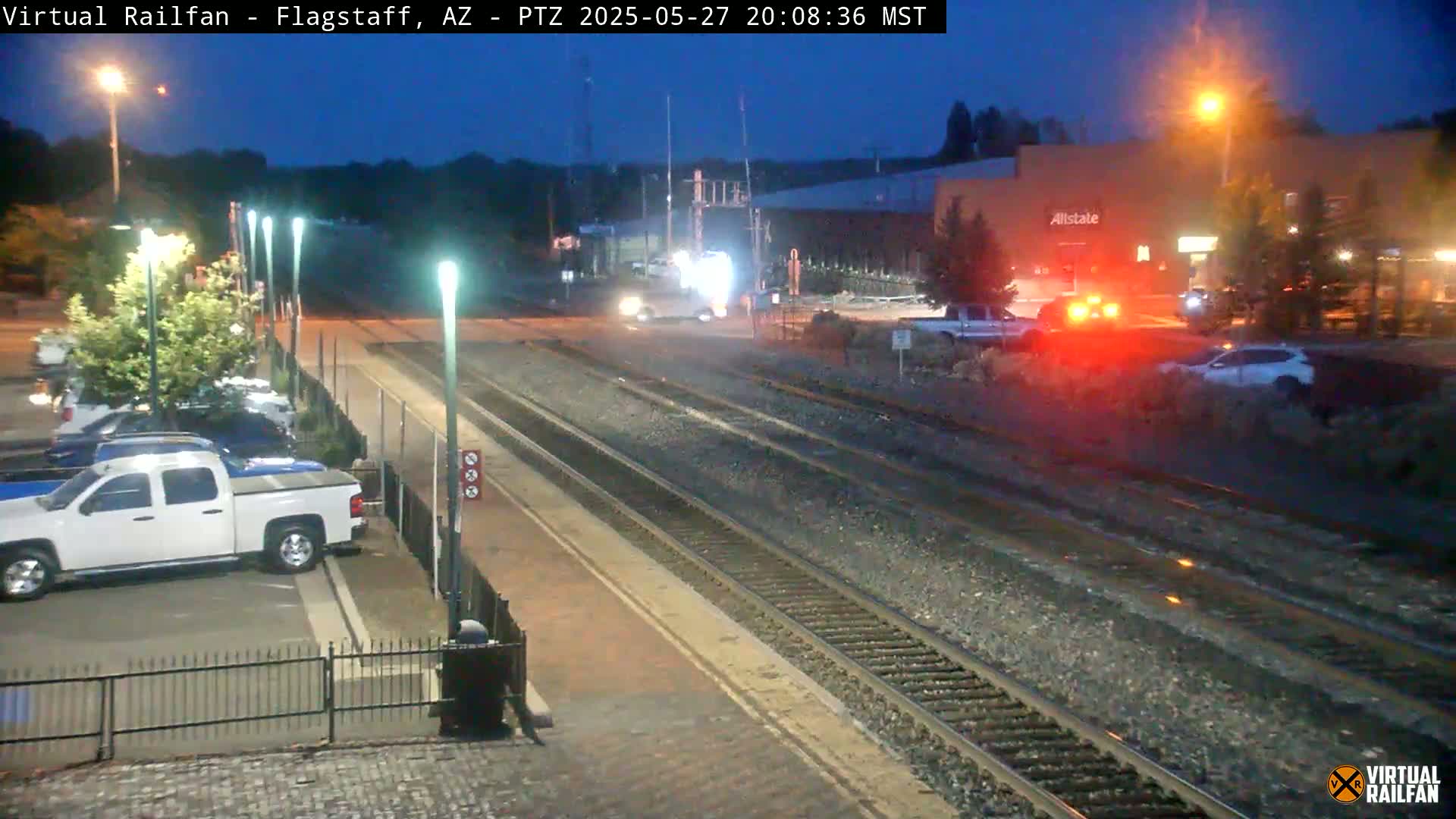 A nighttime view of a train station with multiple train tracks, a parking lot containing several vehicles, and a street with cars driving past a building under a twilight sky.