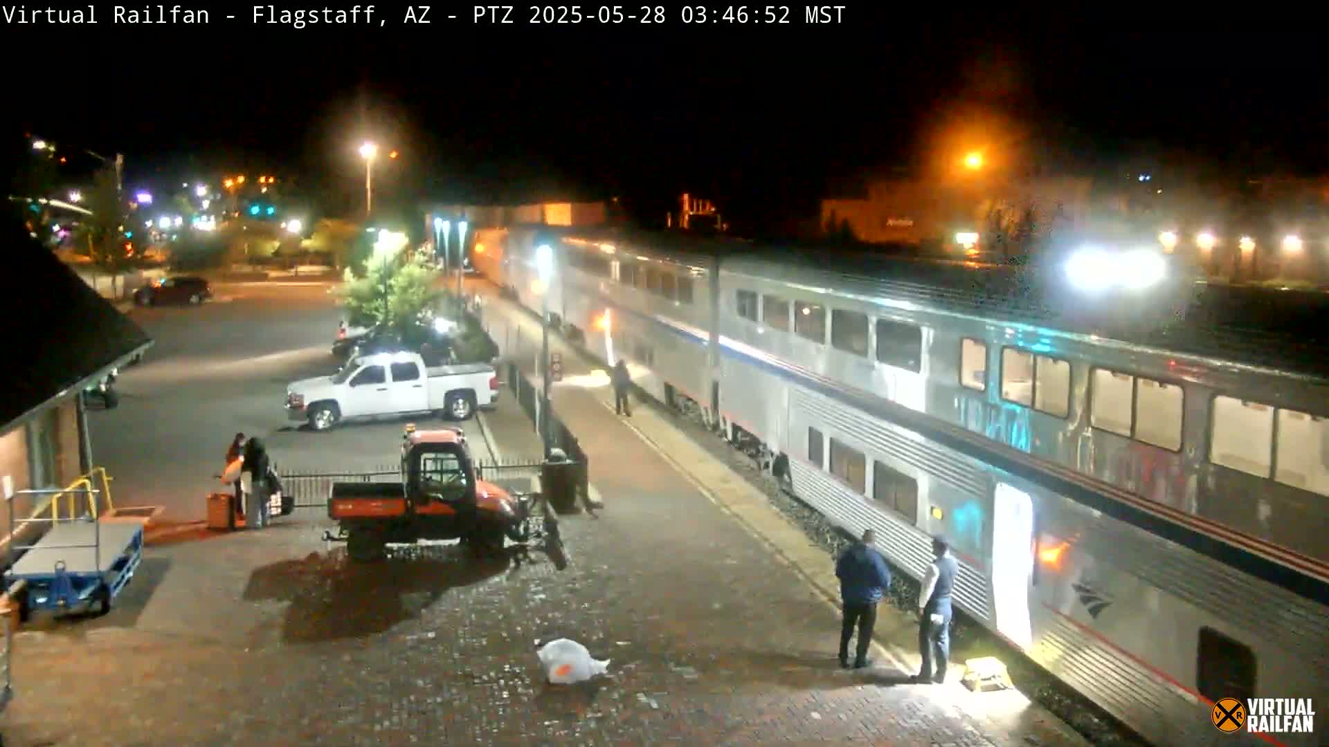At night, a long silver passenger train is stopped at a station platform where several people are standing near it and a few vehicles are parked nearby.