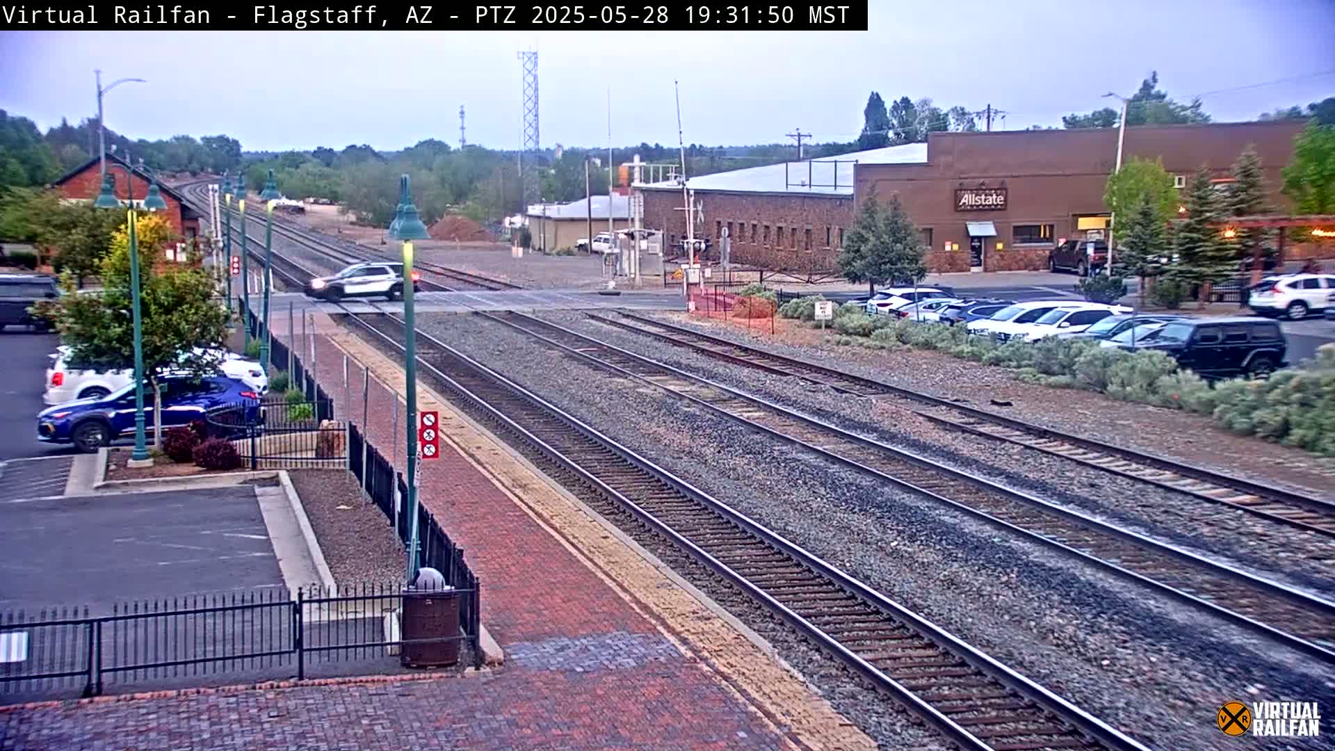 A mostly clear twilight scene shows multiple train tracks intersecting a road with cars parked alongside a building and a vehicle crossing the tracks.