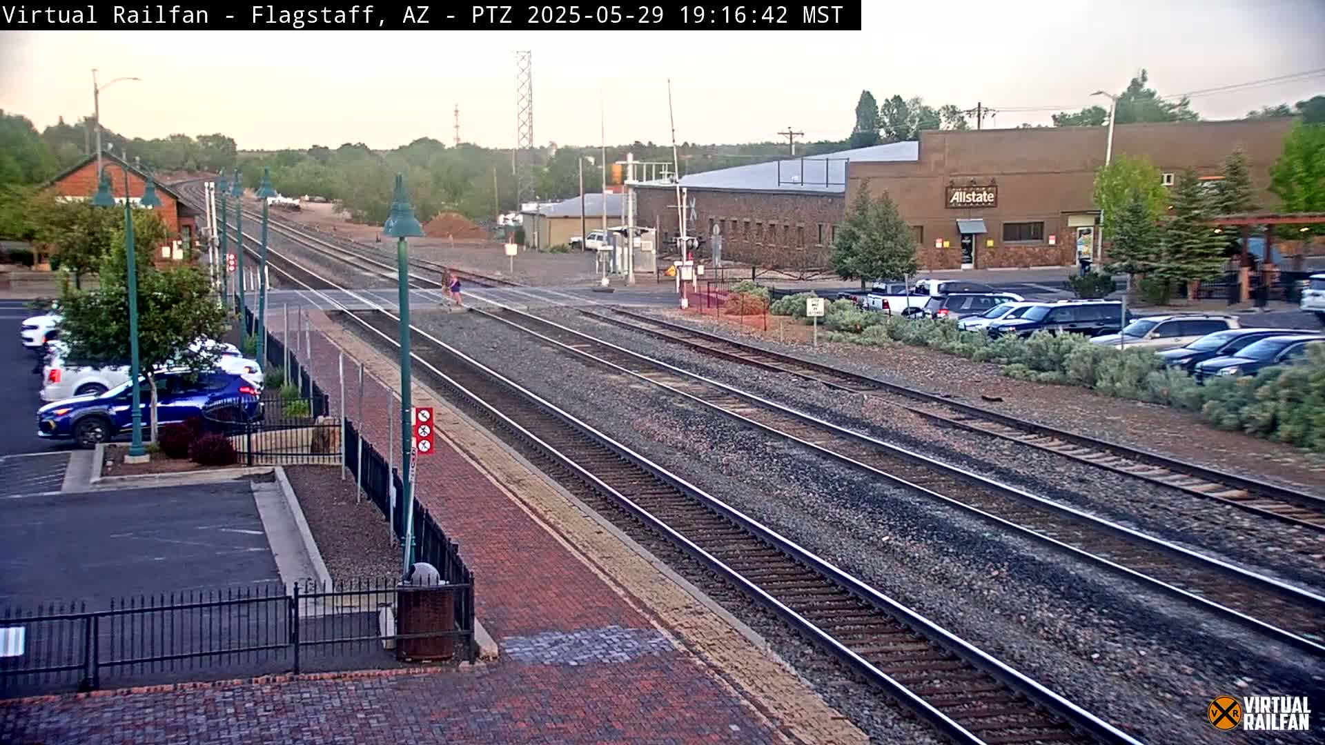 Multiple train tracks run through a town under a partly cloudy sky, with cars parked nearby and buildings visible in the background.