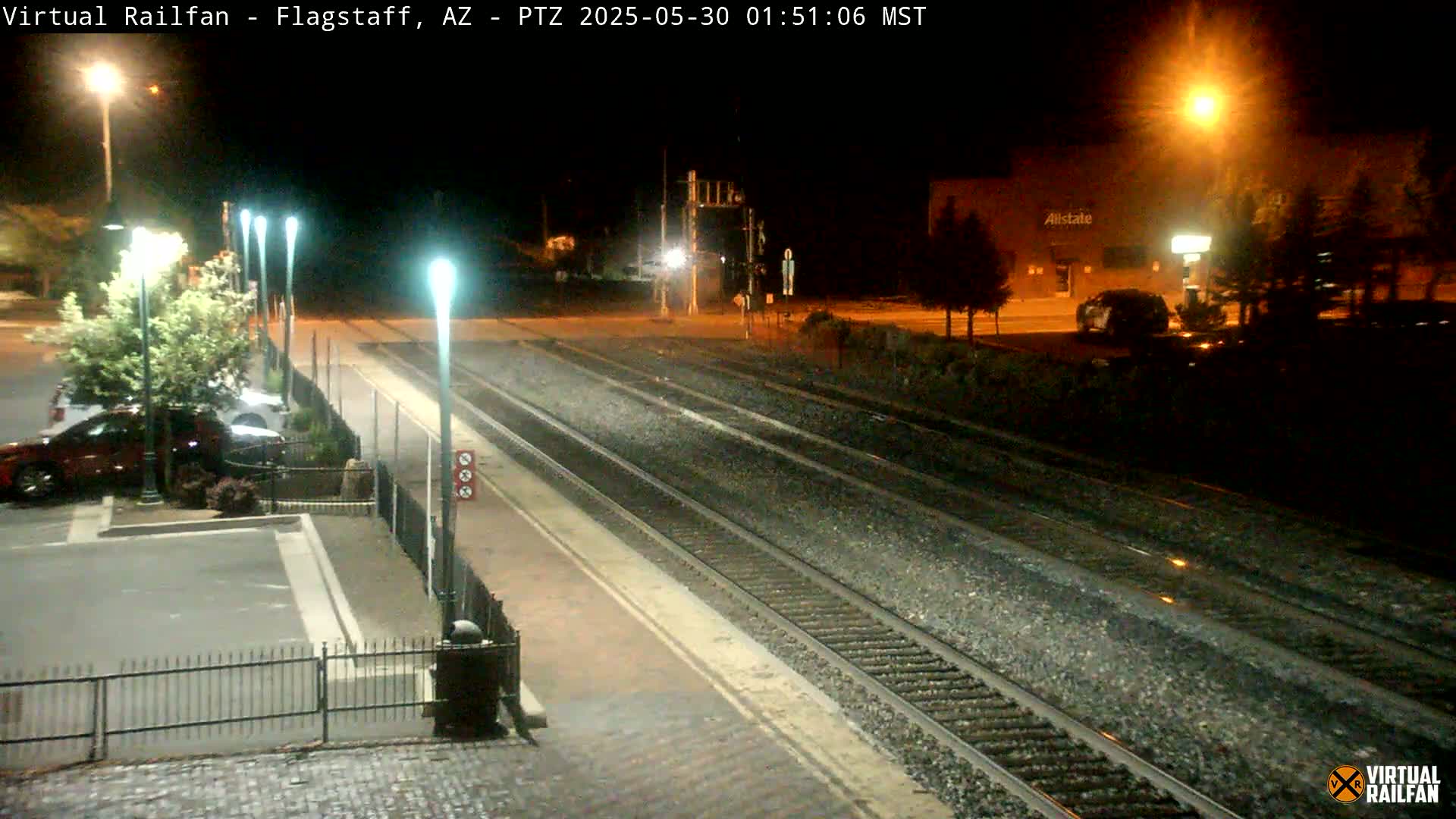 A nighttime view of train tracks at a station, with several cars parked nearby and streetlights illuminating the scene.