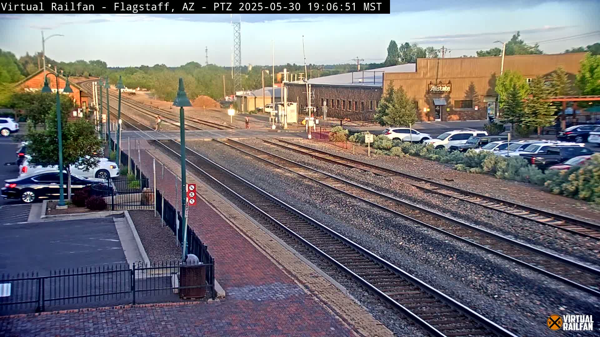 A view of railroad tracks crossing a street in front of a parking lot and buildings under a partly cloudy sky.