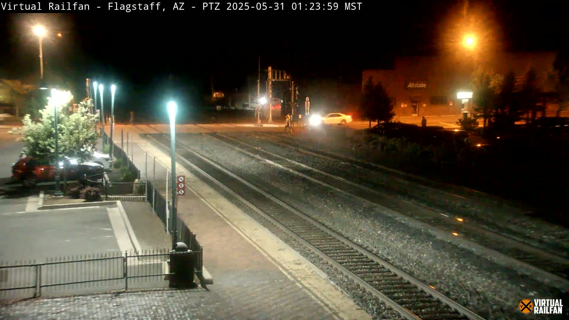 A nighttime view of a train station with multiple train tracks, a parking lot with cars, and streetlights illuminating the area.