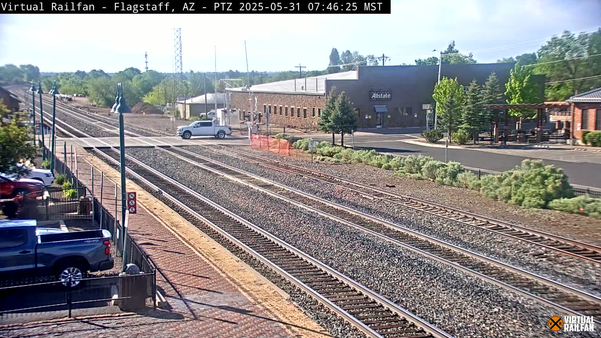A sunny day reveals multiple train tracks intersecting a road near a building with a parking lot, featuring a few vehicles and some greenery.