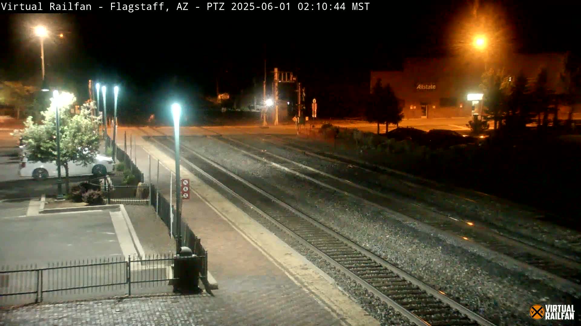 At night, a train station platform is illuminated, showing train tracks extending into the distance, with a parking lot and a building visible in the background under artificial light.