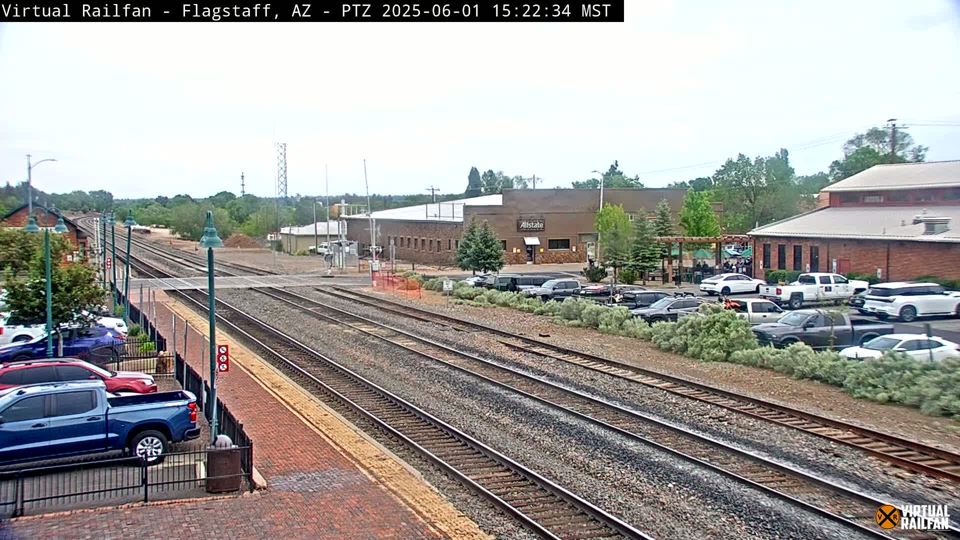 Multiple train tracks run through a town with cars parked along the side, under a partly cloudy sky.