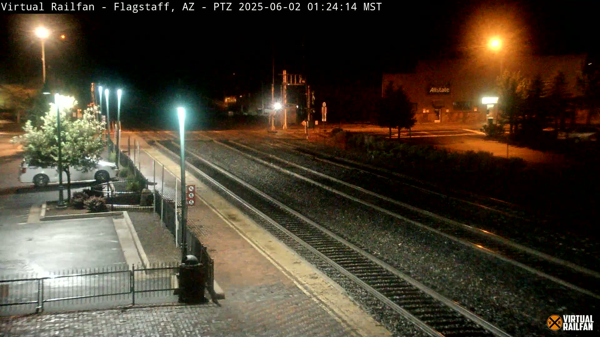 A nighttime view of a train station platform with multiple train tracks, a parked car, and streetlights illuminating the scene.