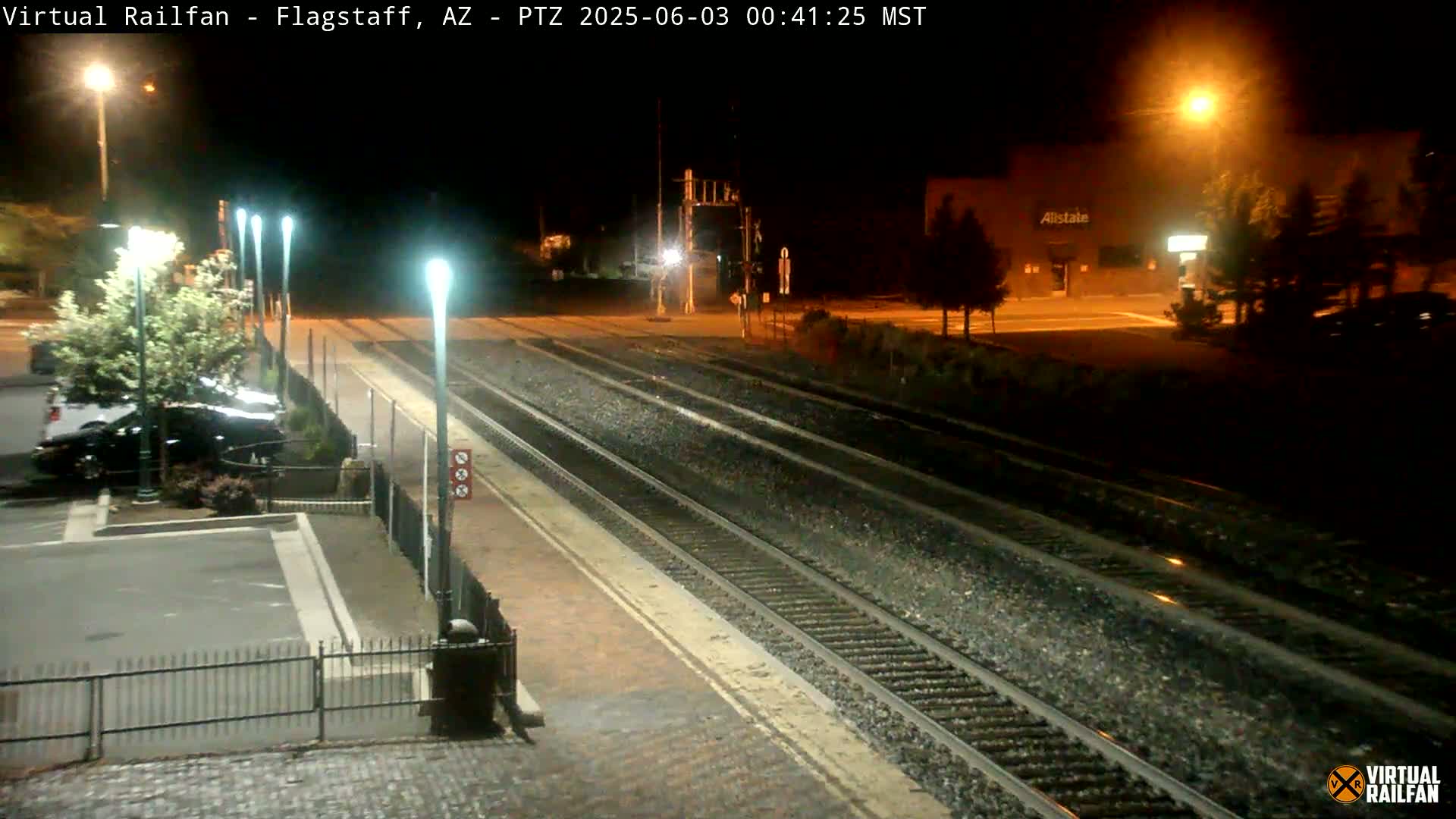 A nighttime view of train tracks at a station, with parked cars visible in the foreground and a building illuminated in the background.