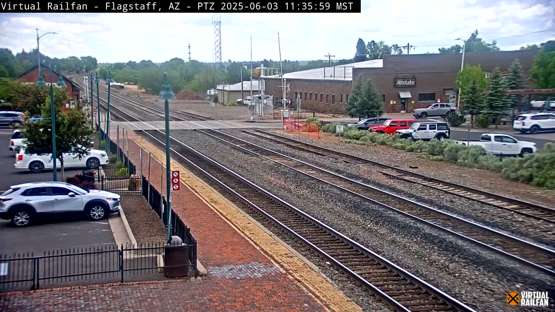 Multiple train tracks stretch into the distance under a partly cloudy sky, passing near a parking lot with cars and a building.
