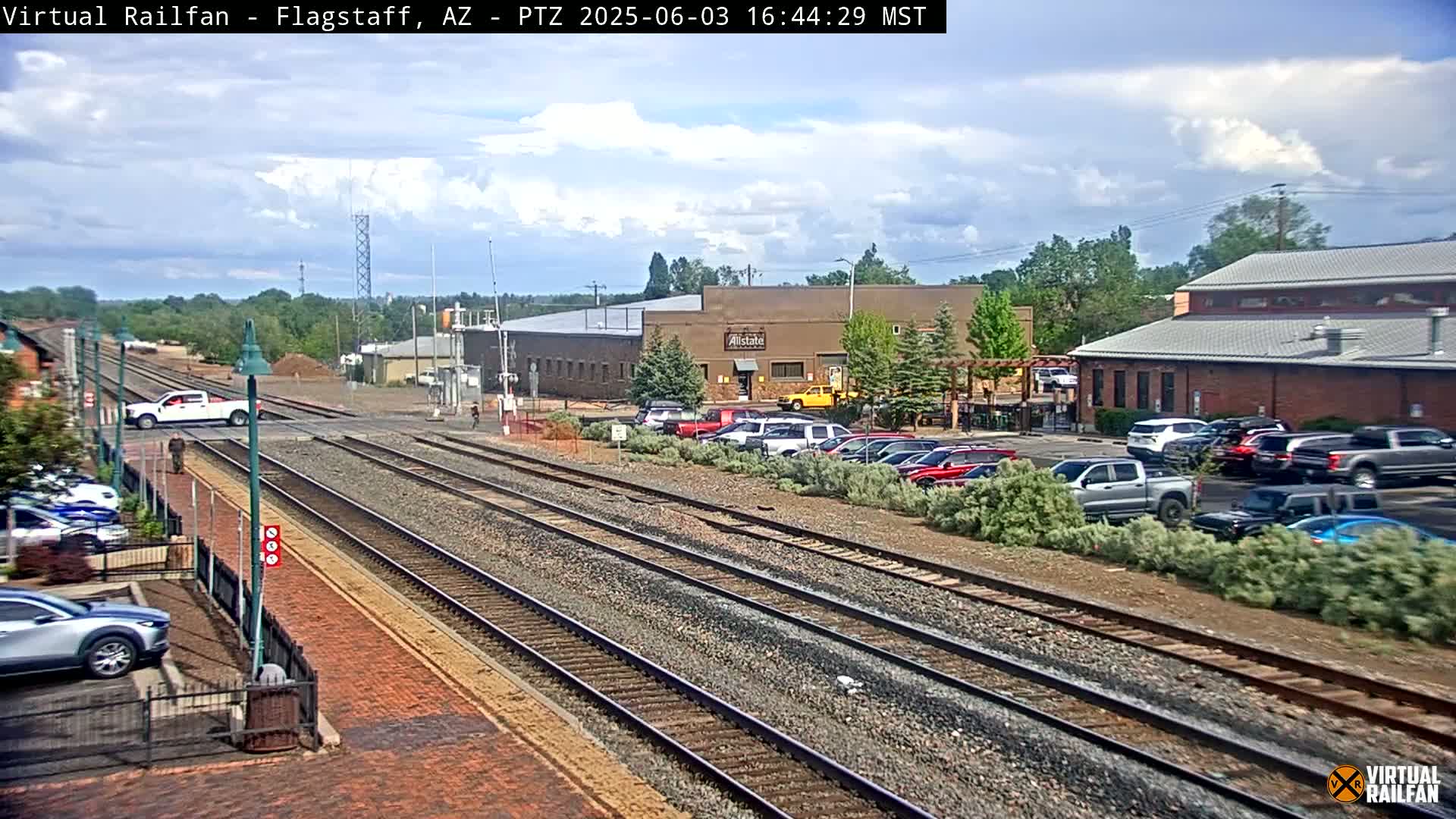 A partly cloudy day shows multiple train tracks intersecting a road with cars parked alongside buildings, including a low brick building and a larger industrial-style structure.