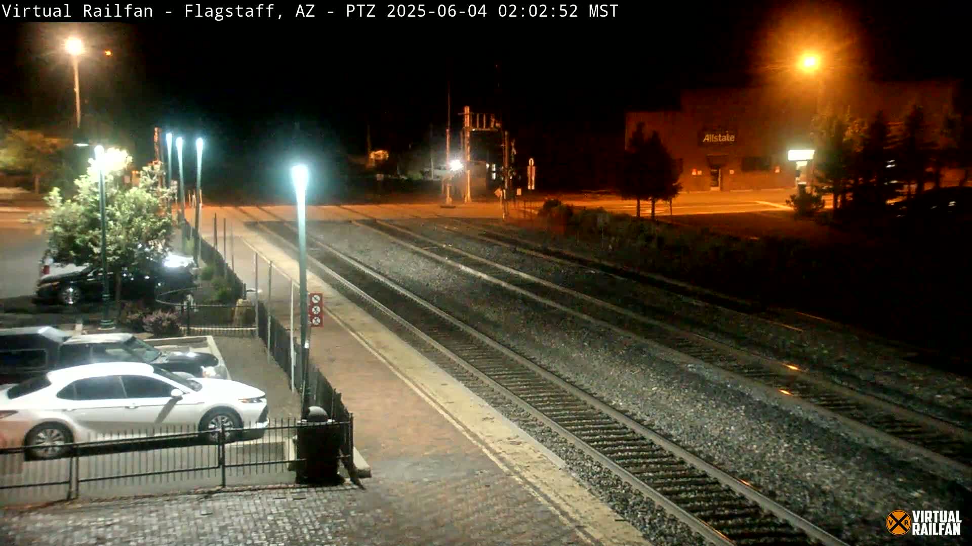 A nighttime view of a train station platform with several sets of train tracks, a parking lot with cars, and a building illuminated in the background.
