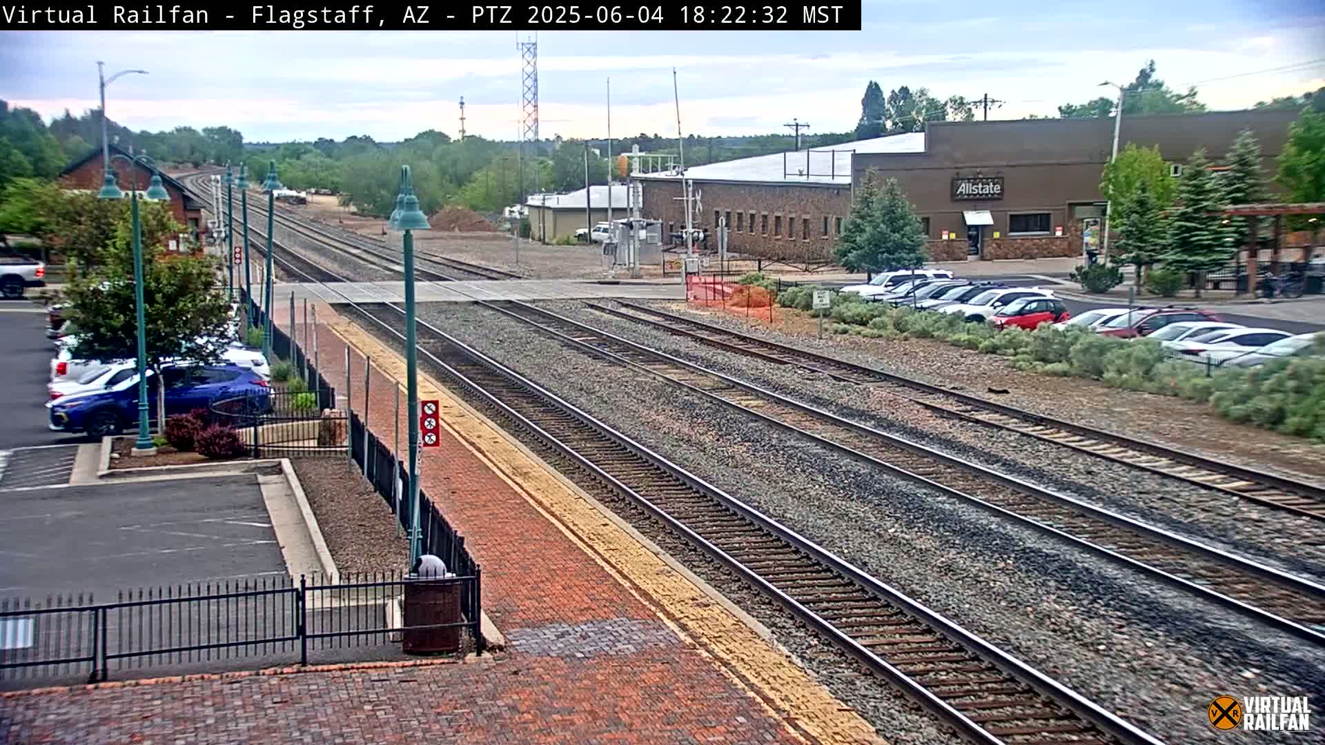 A view of multiple train tracks extending into the distance, alongside a brick paved area, parked cars, and buildings under an overcast sky.