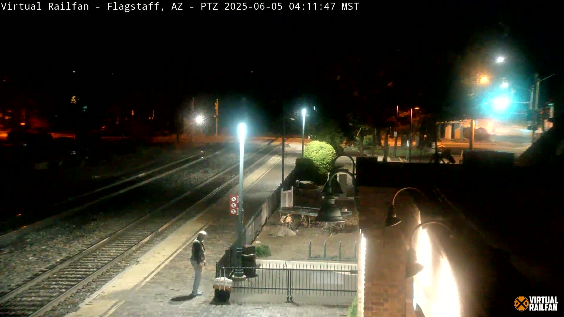 A nighttime view of a train station platform with a person standing near a bell, under illuminated streetlights.