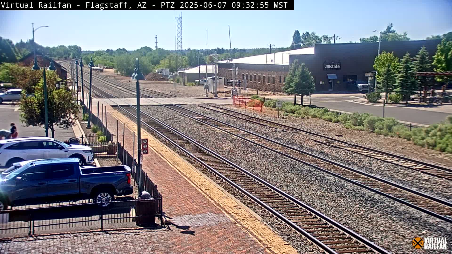 A sunny outdoor view of train tracks, a parking lot with a few vehicles, and buildings in the distance.