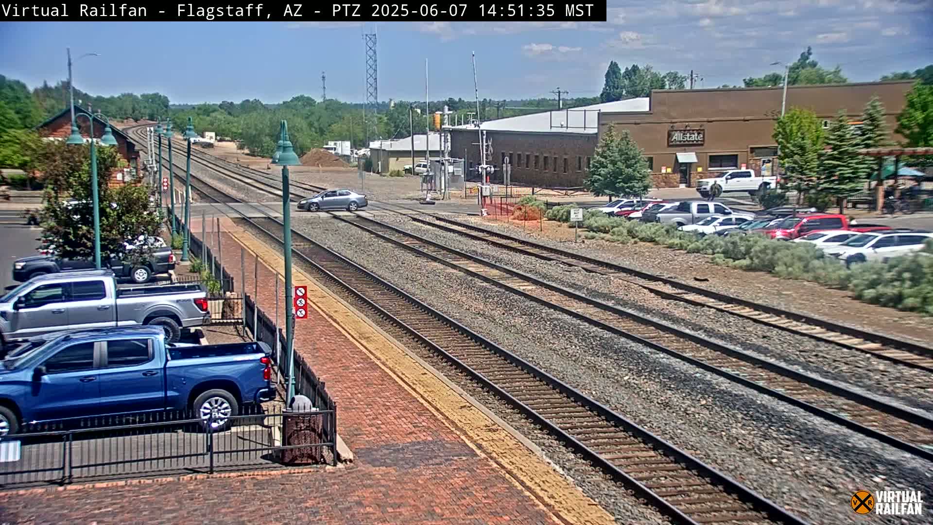 A sunny day shows multiple sets of train tracks intersecting a road, with cars parked nearby and a few cars driving across the crossing.