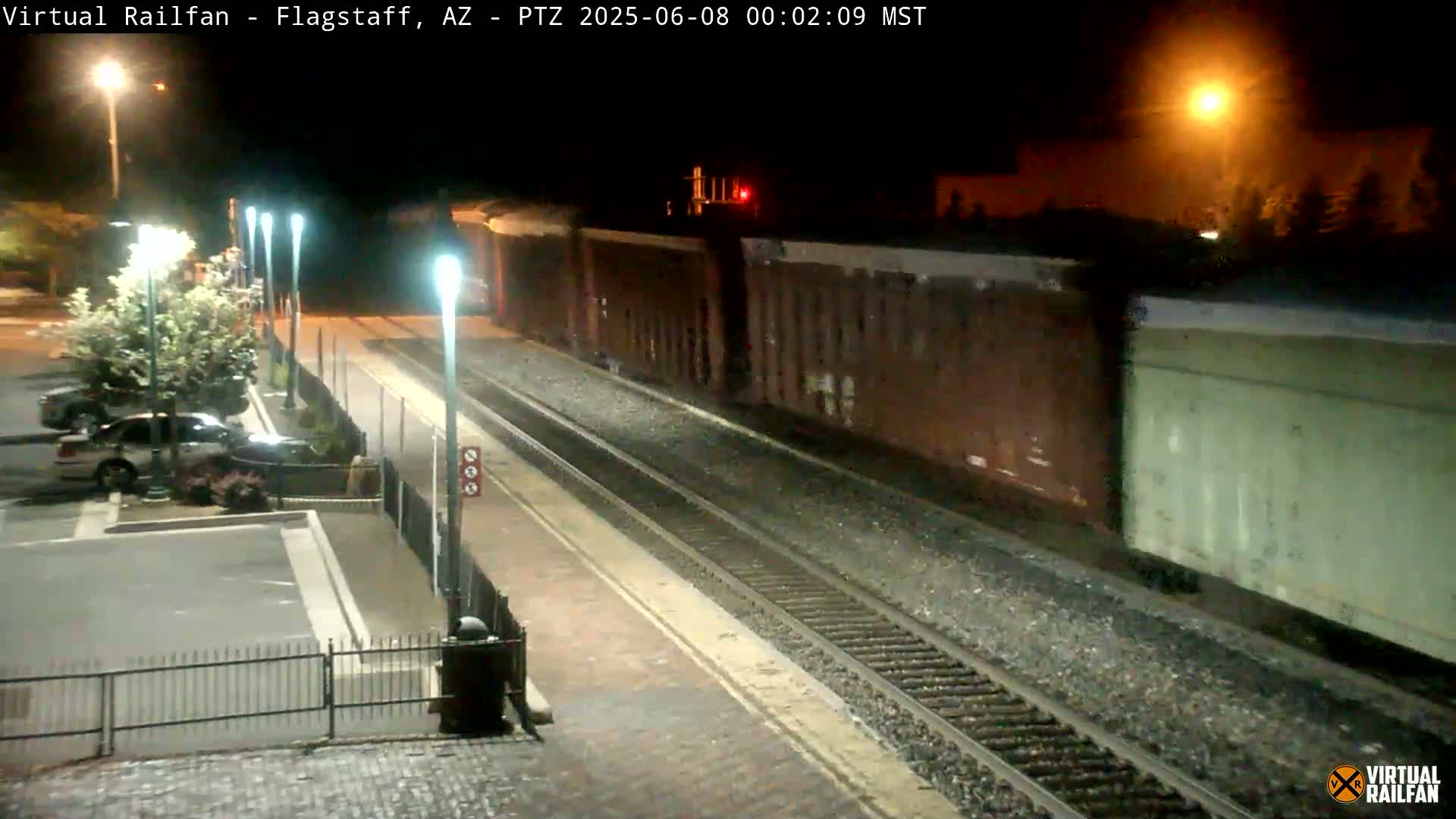 A long freight train passes by a nighttime train station with cars parked in a nearby lot.