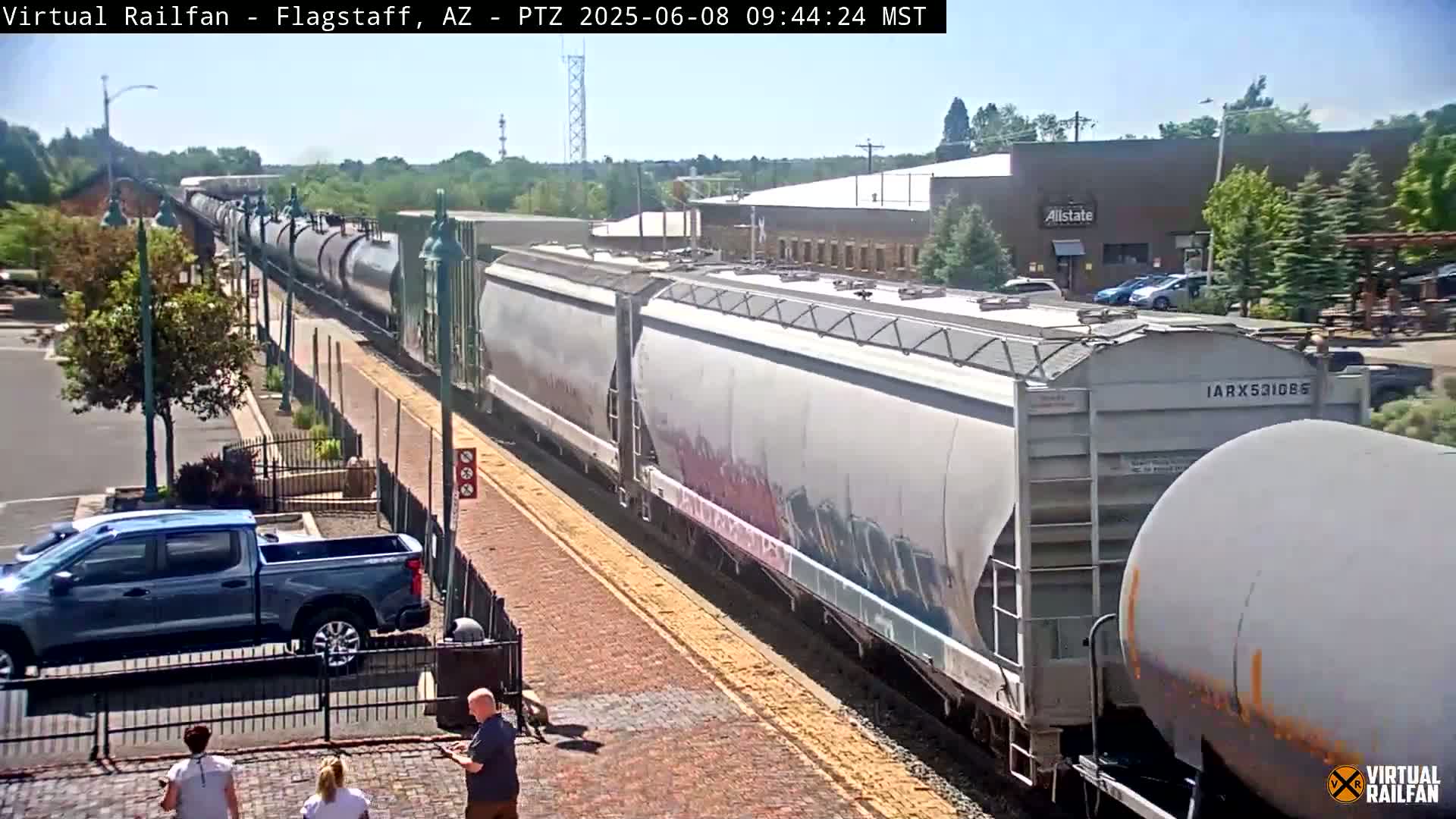 A long freight train passes by on a sunny day next to a street with parked cars and pedestrians.
