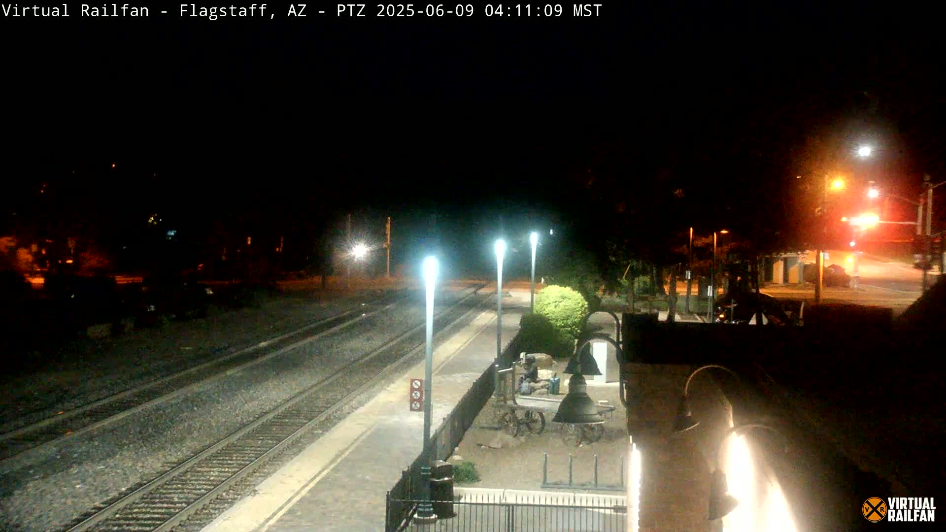A nighttime view of a train station platform with train tracks, several lights, and a large bell in the foreground.
