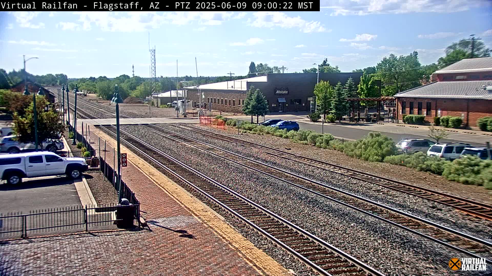 A sunny day view of railroad tracks at a train station with several cars parked nearby, and buildings in the background.