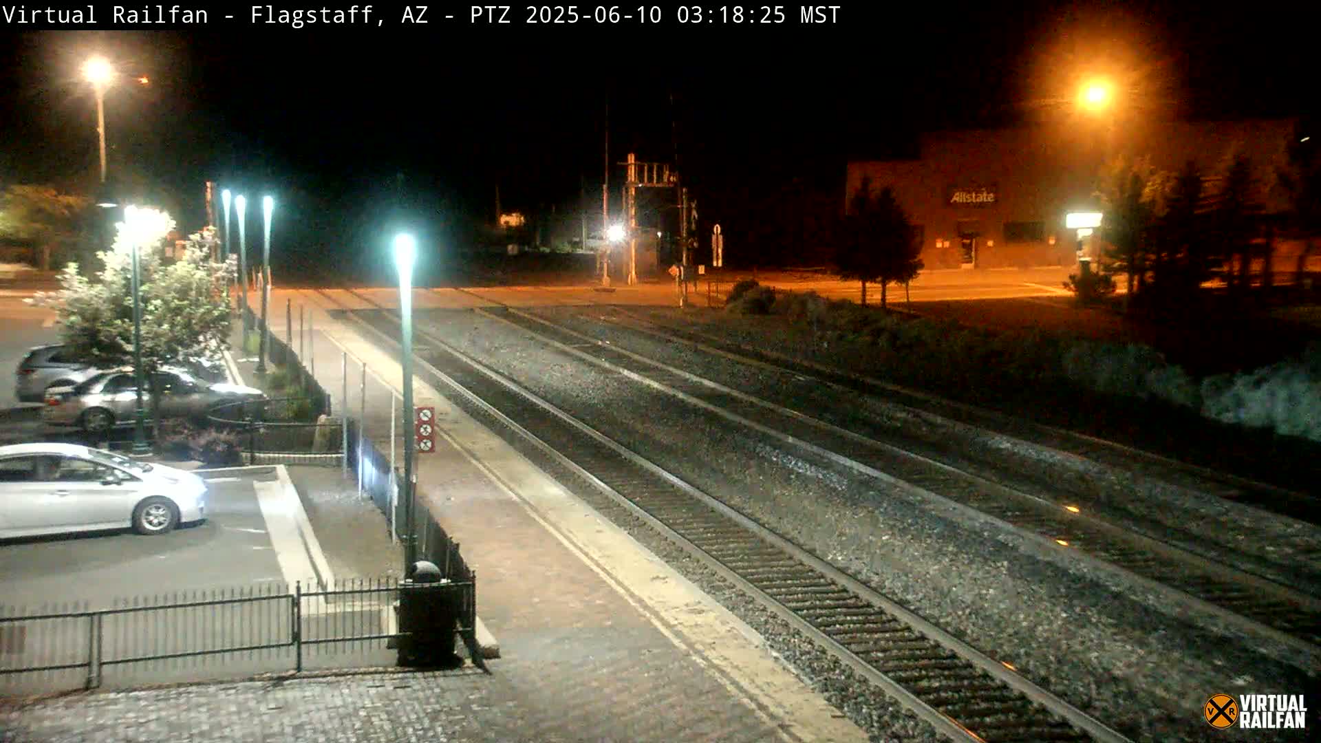 A nighttime view of train tracks at a station with cars parked nearby, illuminated by streetlights.