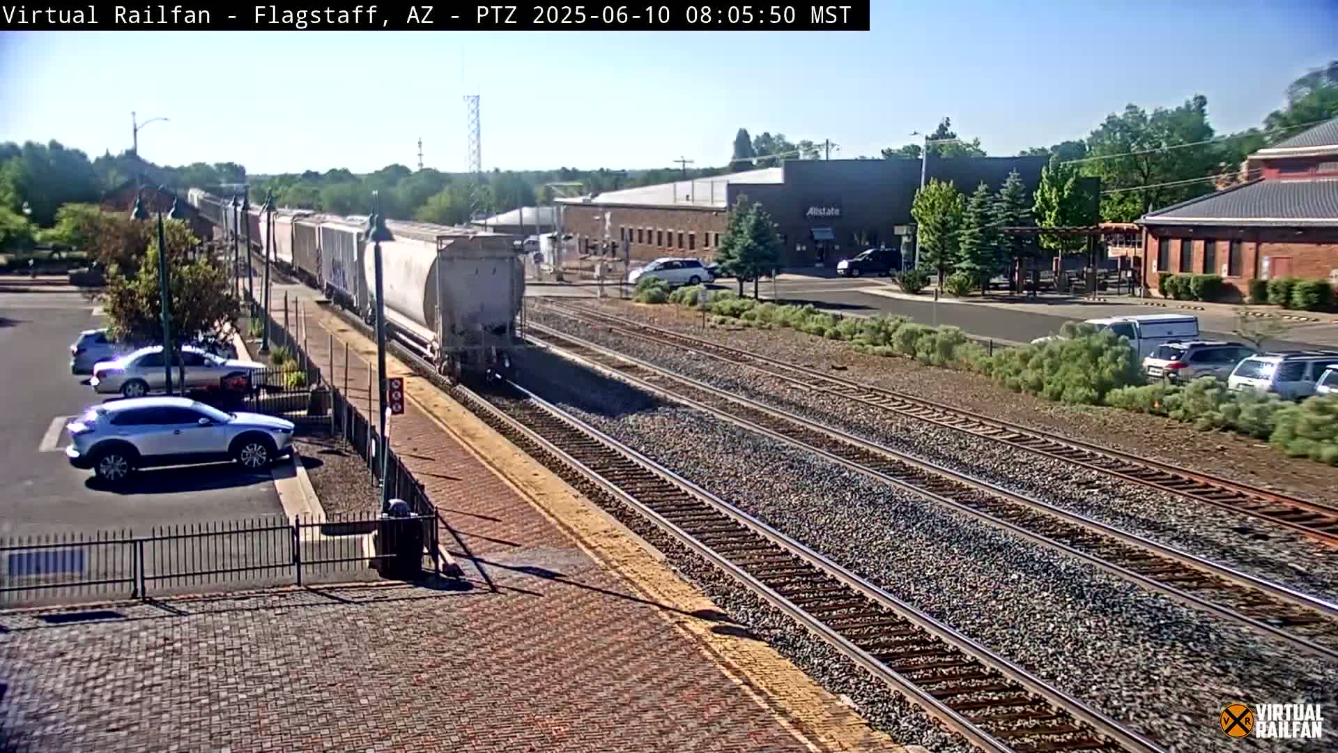 A long freight train passes by a railway station on a sunny day, with several cars parked in a nearby lot.