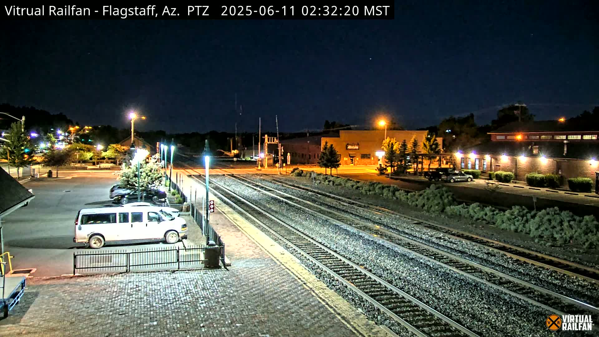 A nighttime view of a train station with several sets of tracks, a parking lot with a white van, and buildings under a clear, dark sky.