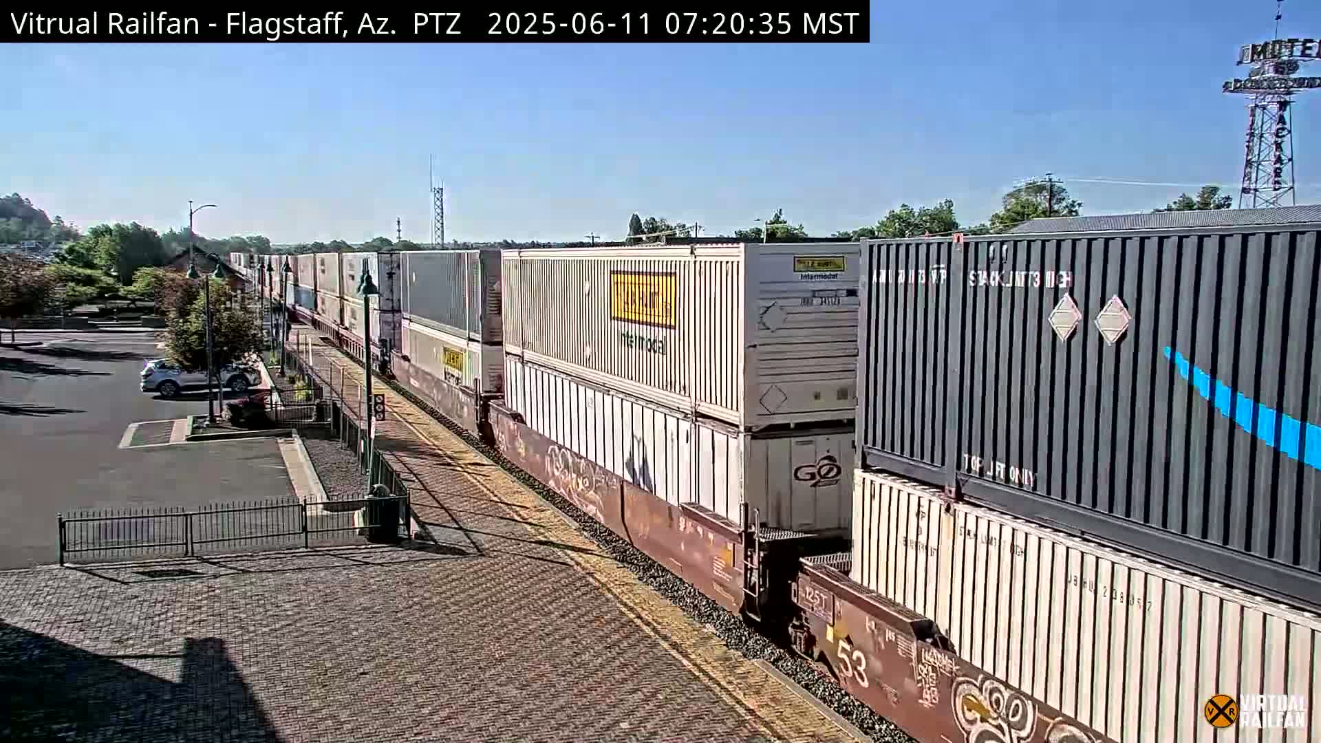 A long freight train carrying multiple shipping containers moves along a railway track under a clear blue sky.
