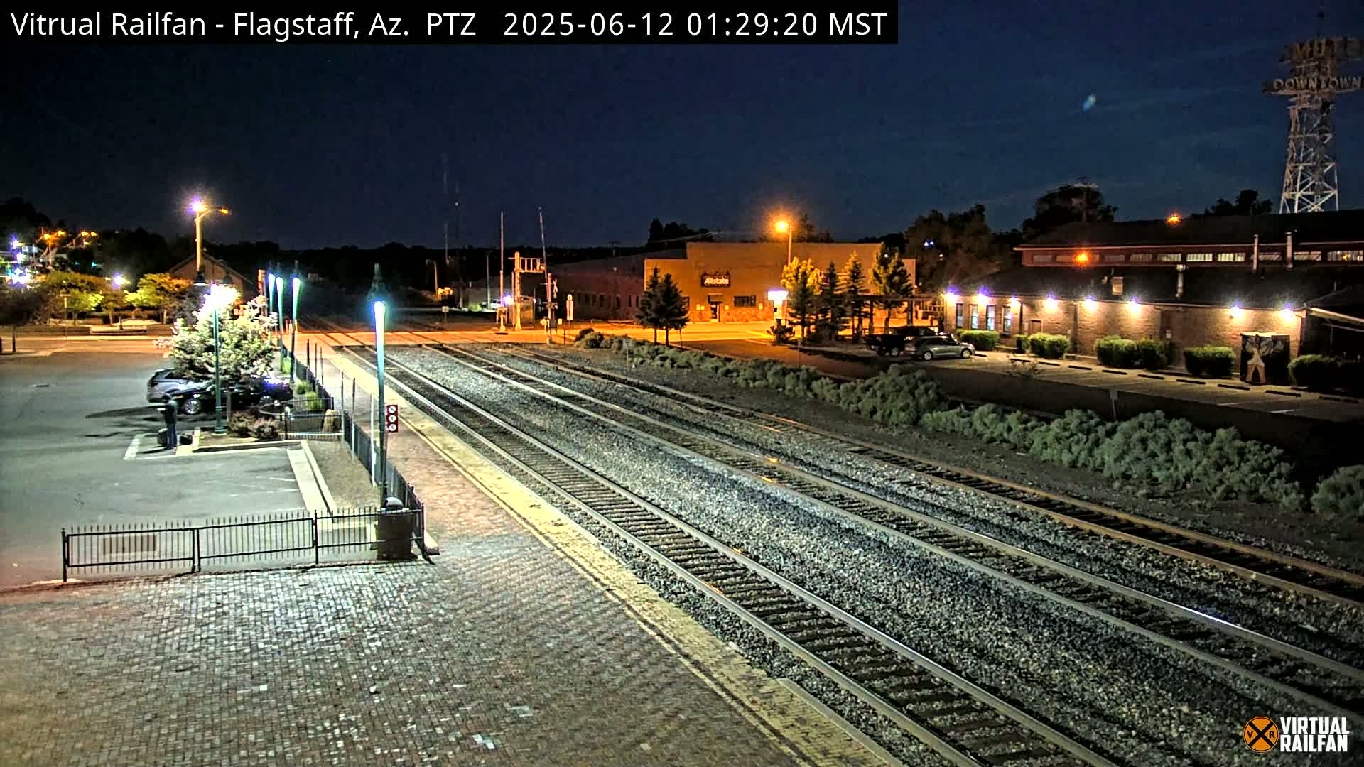 A nighttime view of a train station platform and tracks, with a parking lot and buildings visible in the background under a clear, dark sky.
