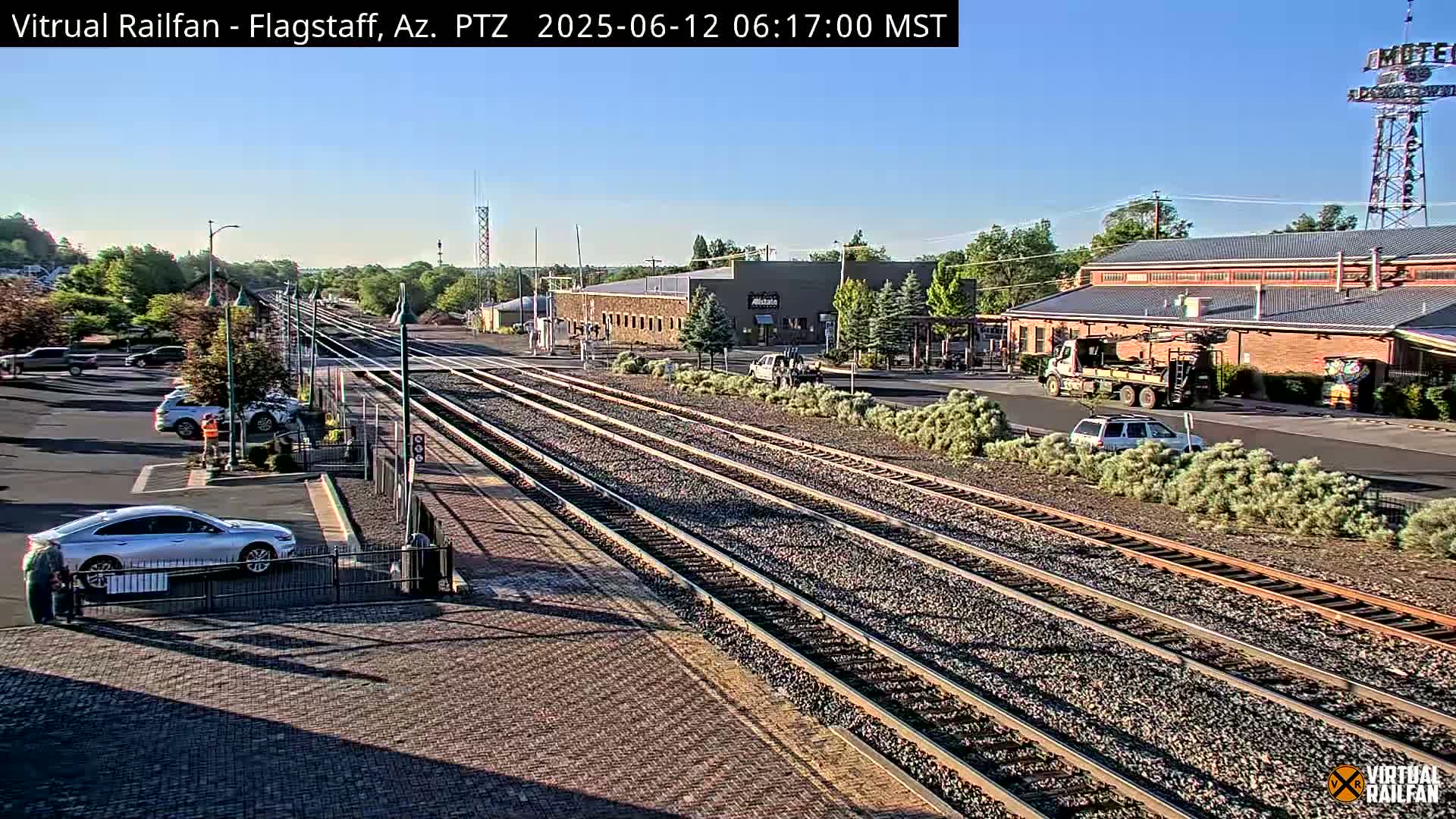 Multiple train tracks stretch toward a brick building and parking lot under a clear, sunny sky.