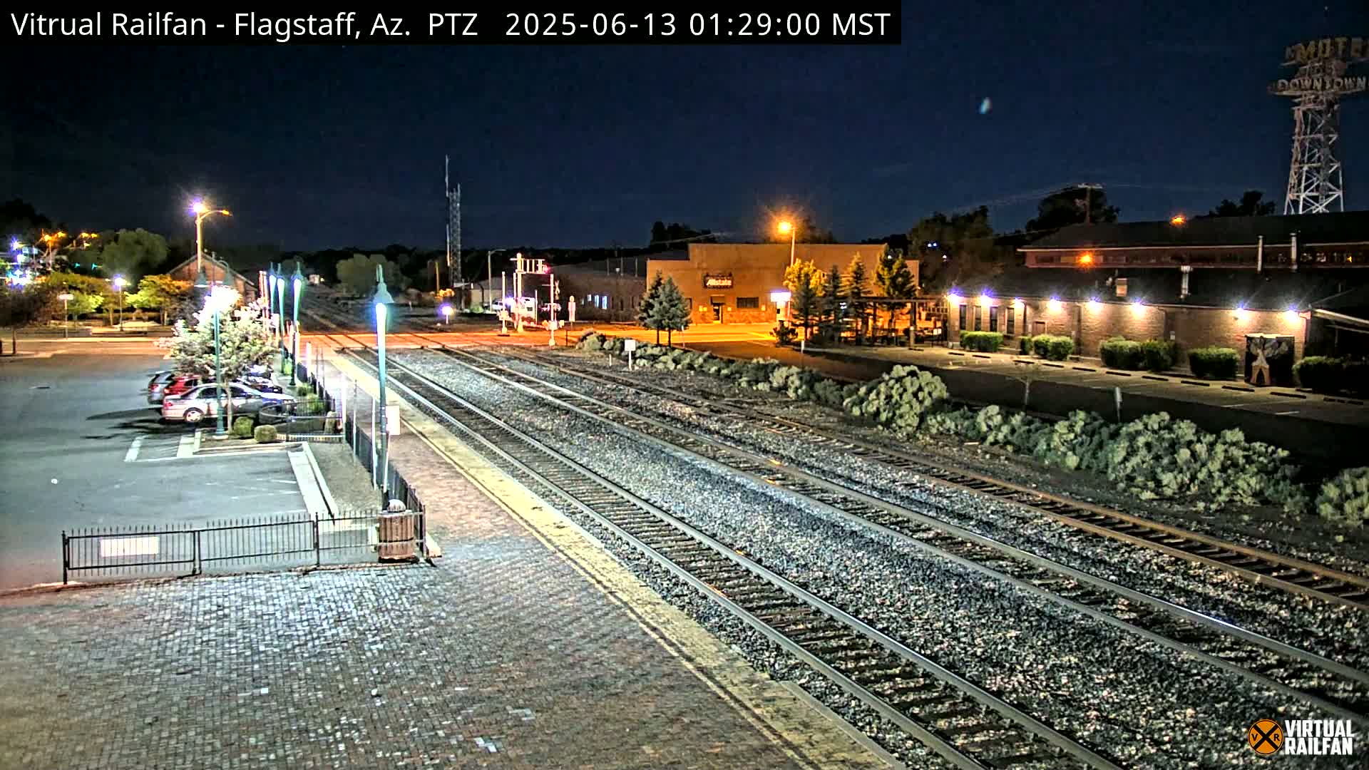 A nighttime view of train tracks extending from a brick paved area towards buildings under a clear, dark sky.