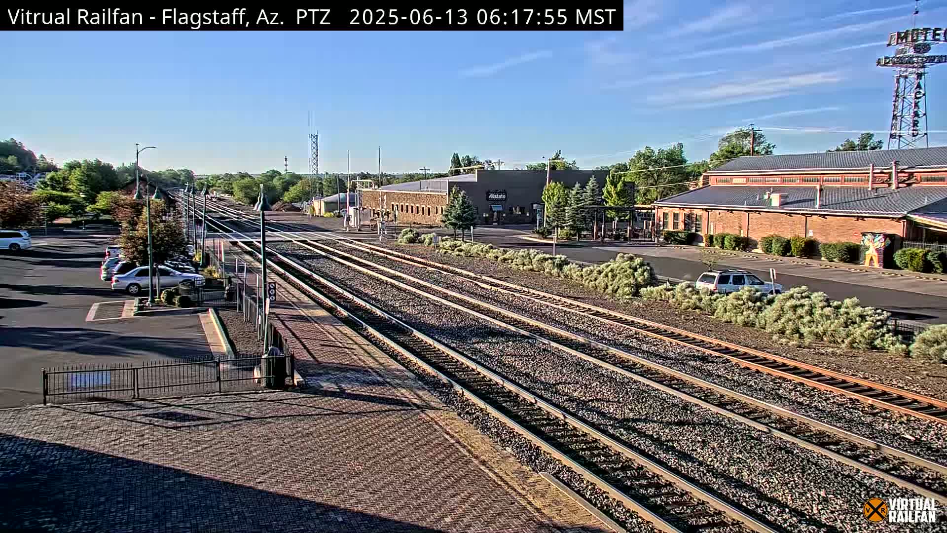 A sunny day shows multiple train tracks extending from a parking lot with several cars, past industrial buildings, and toward a clear sky.