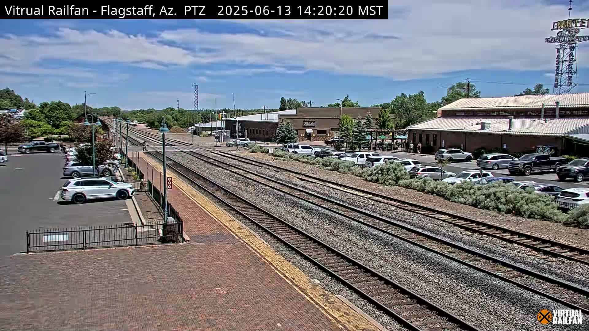 A sunny day at a train station with multiple tracks, a parking lot full of cars, and several buildings in the background.