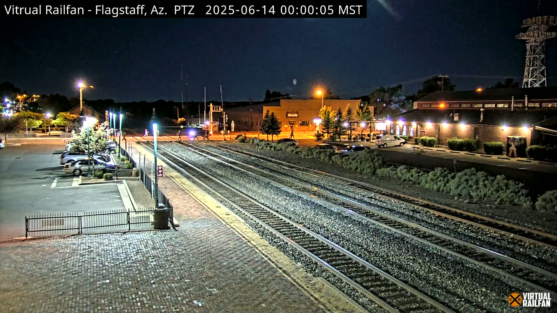 A nighttime view of a train station with multiple train tracks, a parking lot with several cars, and buildings under a clear, dark sky.