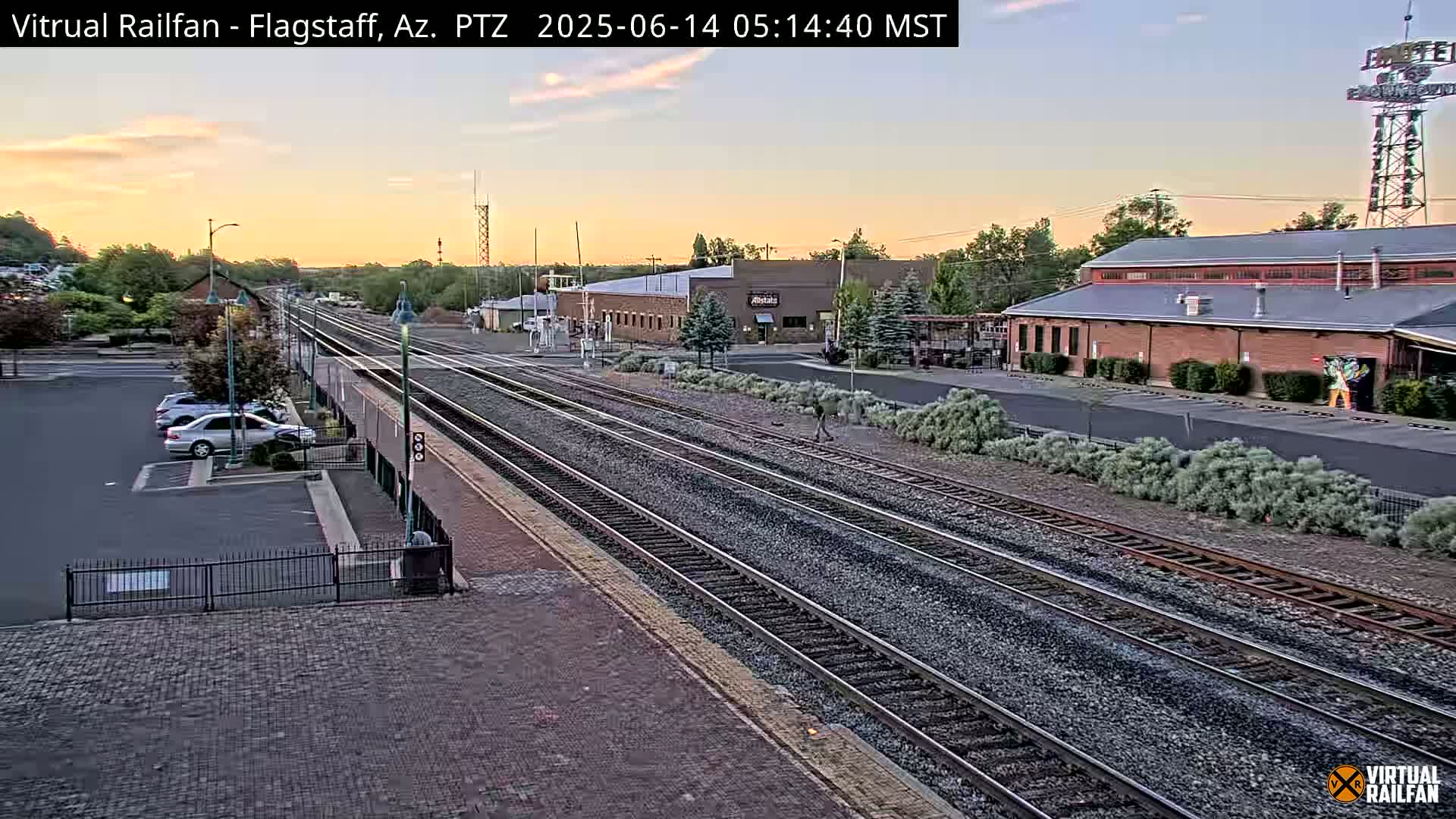 A sunrise illuminates a train station with multiple tracks, a parking lot with several cars, and low buildings under a clear sky.