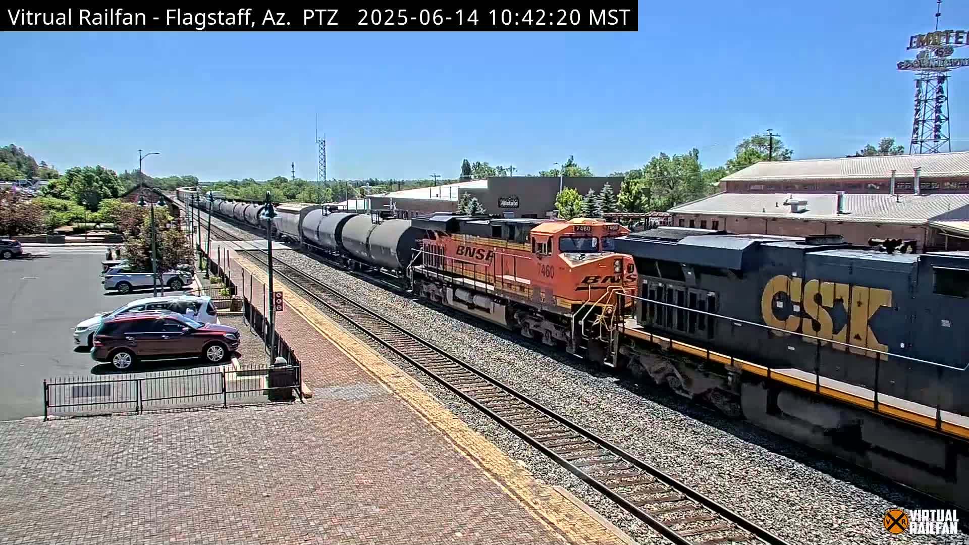 A long freight train, led by an orange locomotive, passes through a train station on a sunny day.
