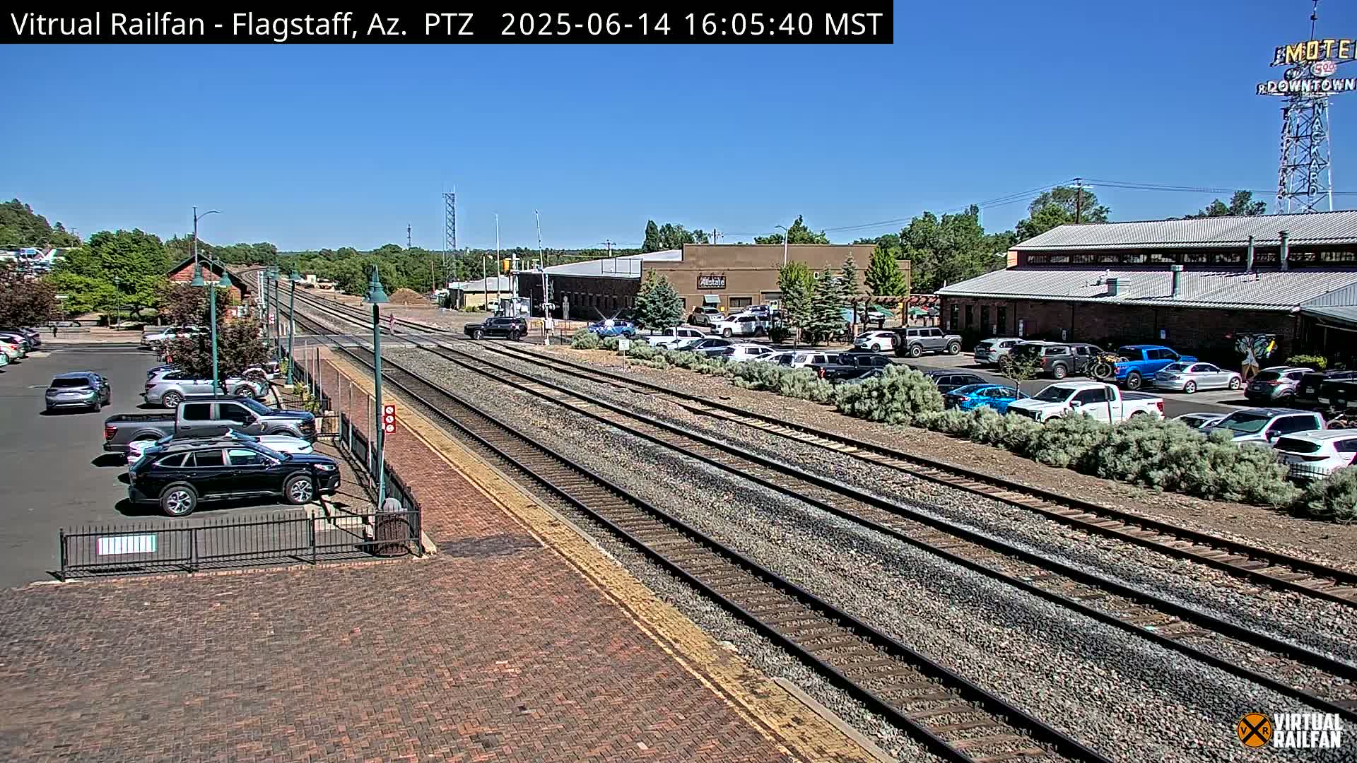 A long freight train, led by an orange locomotive, passes through a train station on a sunny day.