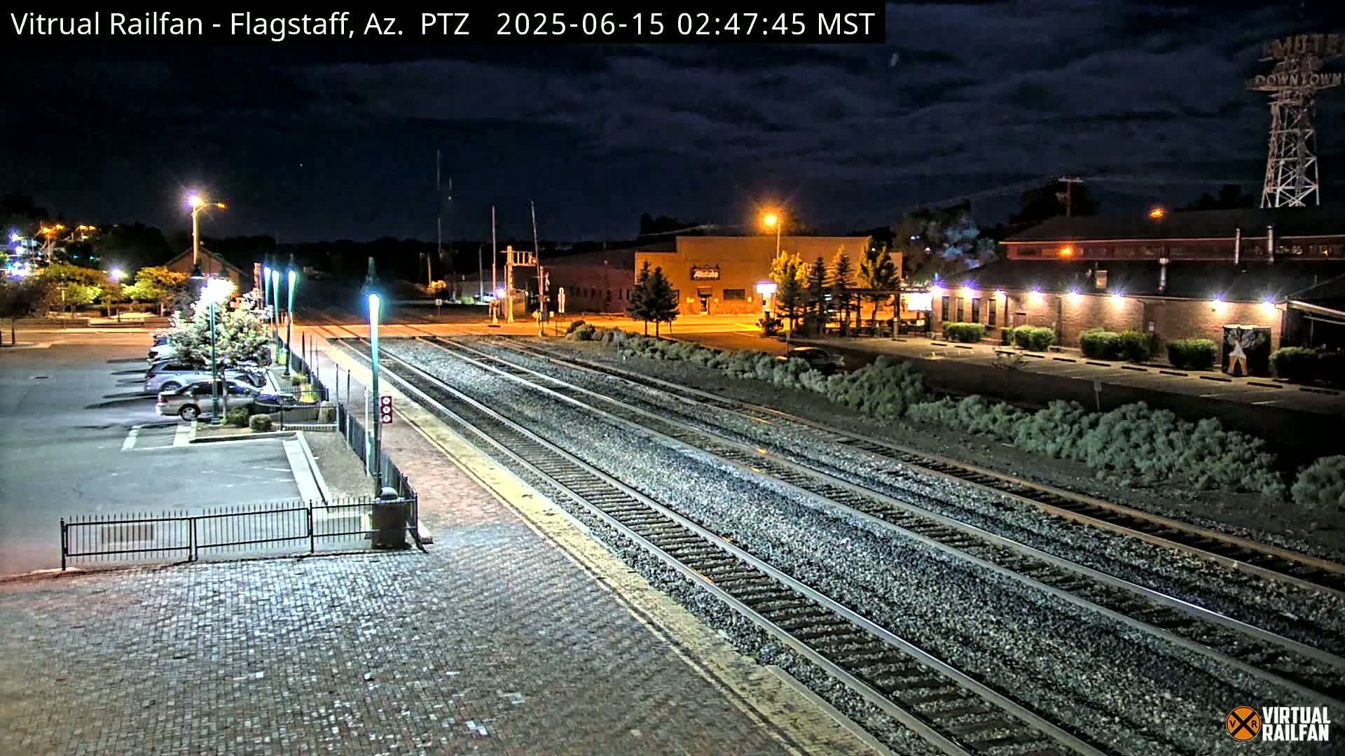 A nighttime view of a train station with multiple sets of tracks, a parking lot with cars, and buildings under a partly cloudy sky.