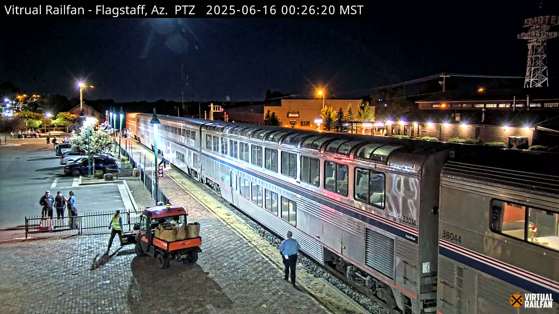 At night, a long silver passenger train is at a station, with several people standing nearby and a small vehicle loading items onto it.