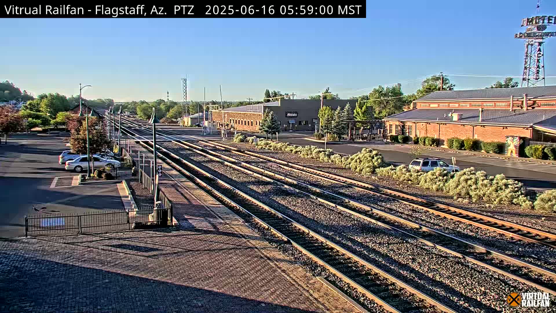 Under a clear, sunny sky, multiple train tracks extend from a paved parking lot with several cars, past low buildings and shrubbery, towards a distant horizon.