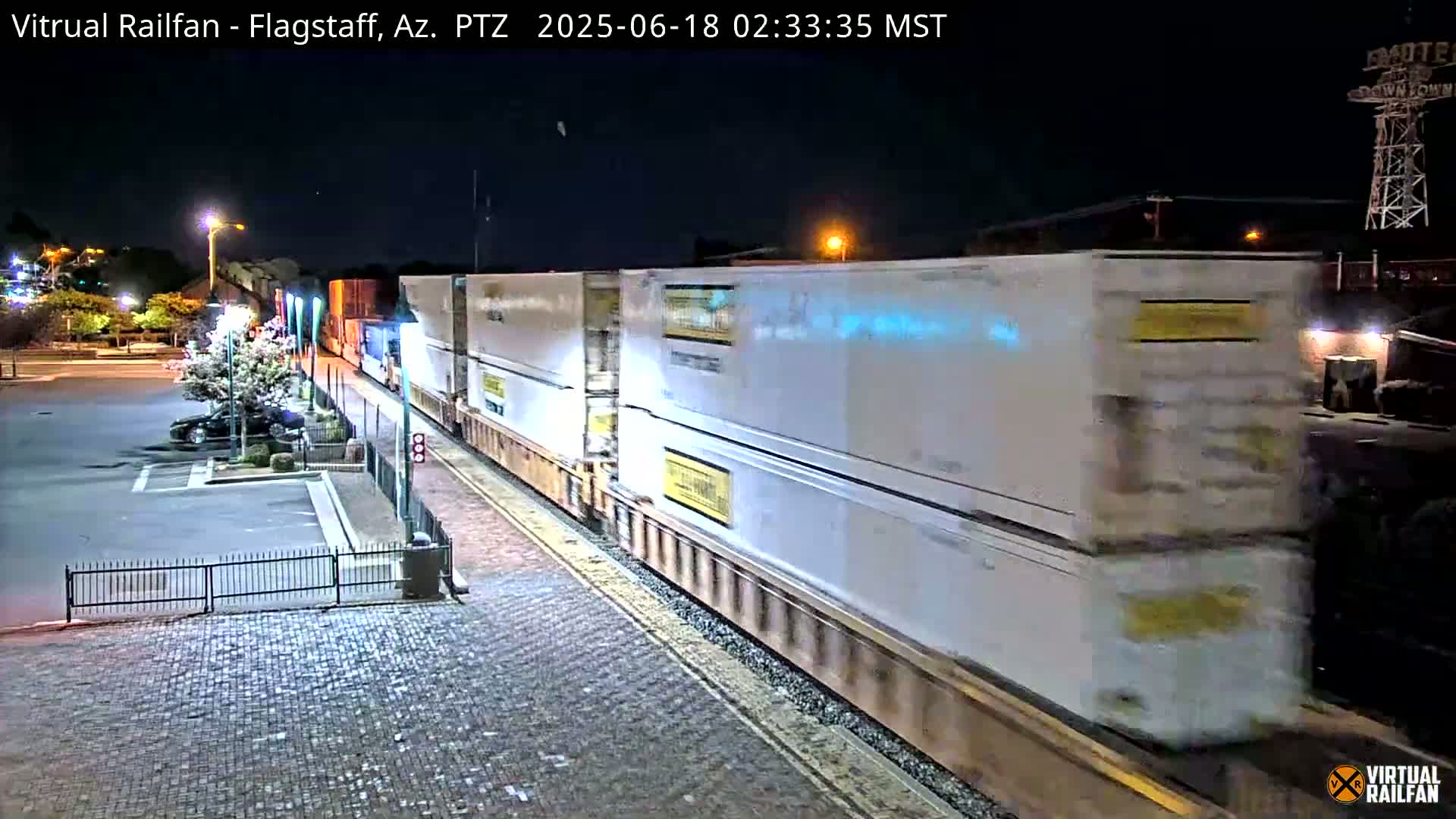 A long freight train carrying numerous white shipping containers moves slowly through a station at night.