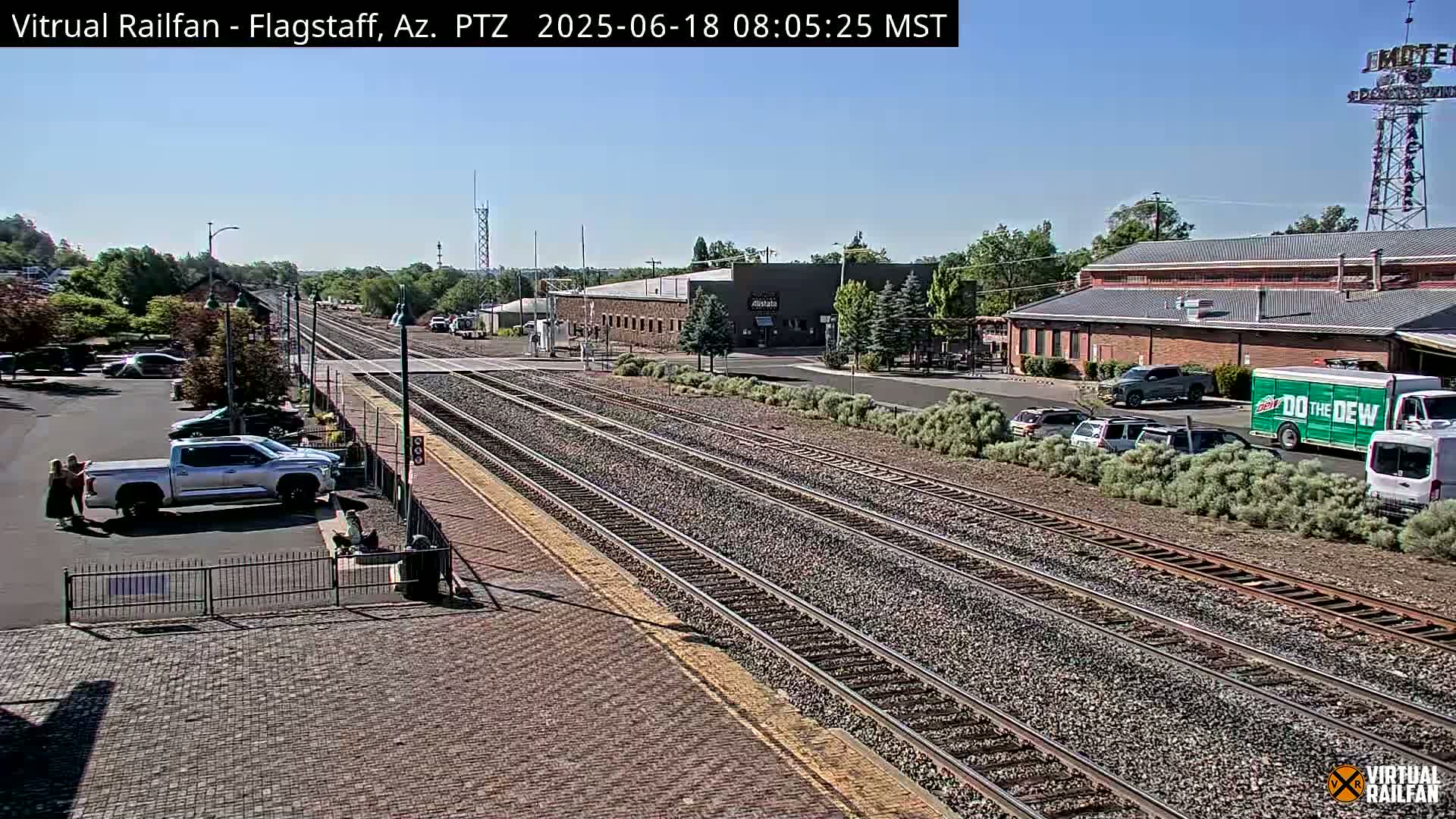 A sunny day reveals multiple train tracks running through a town with parked cars and buildings, including a Mountain Dew truck.