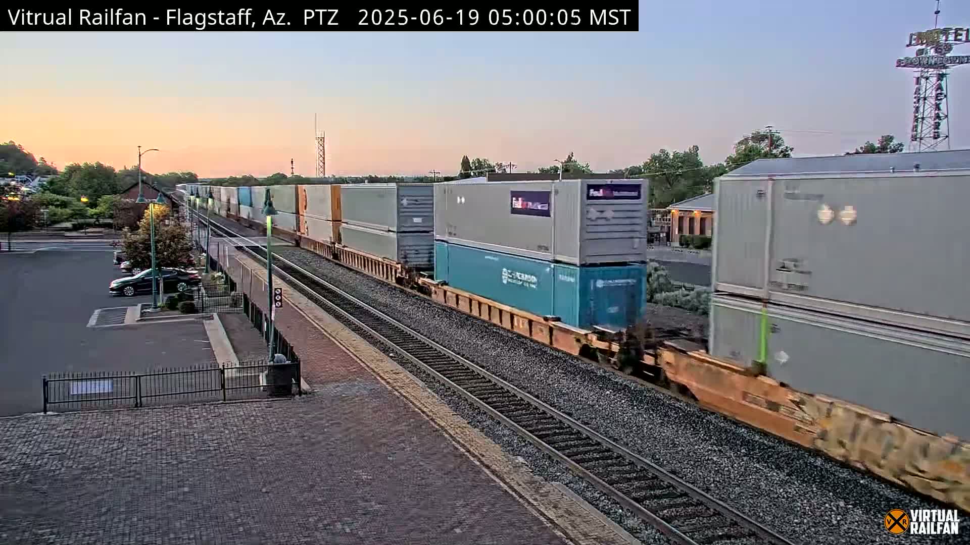 A long freight train carrying multiple shipping containers passes by a train station during a twilight sunrise.
