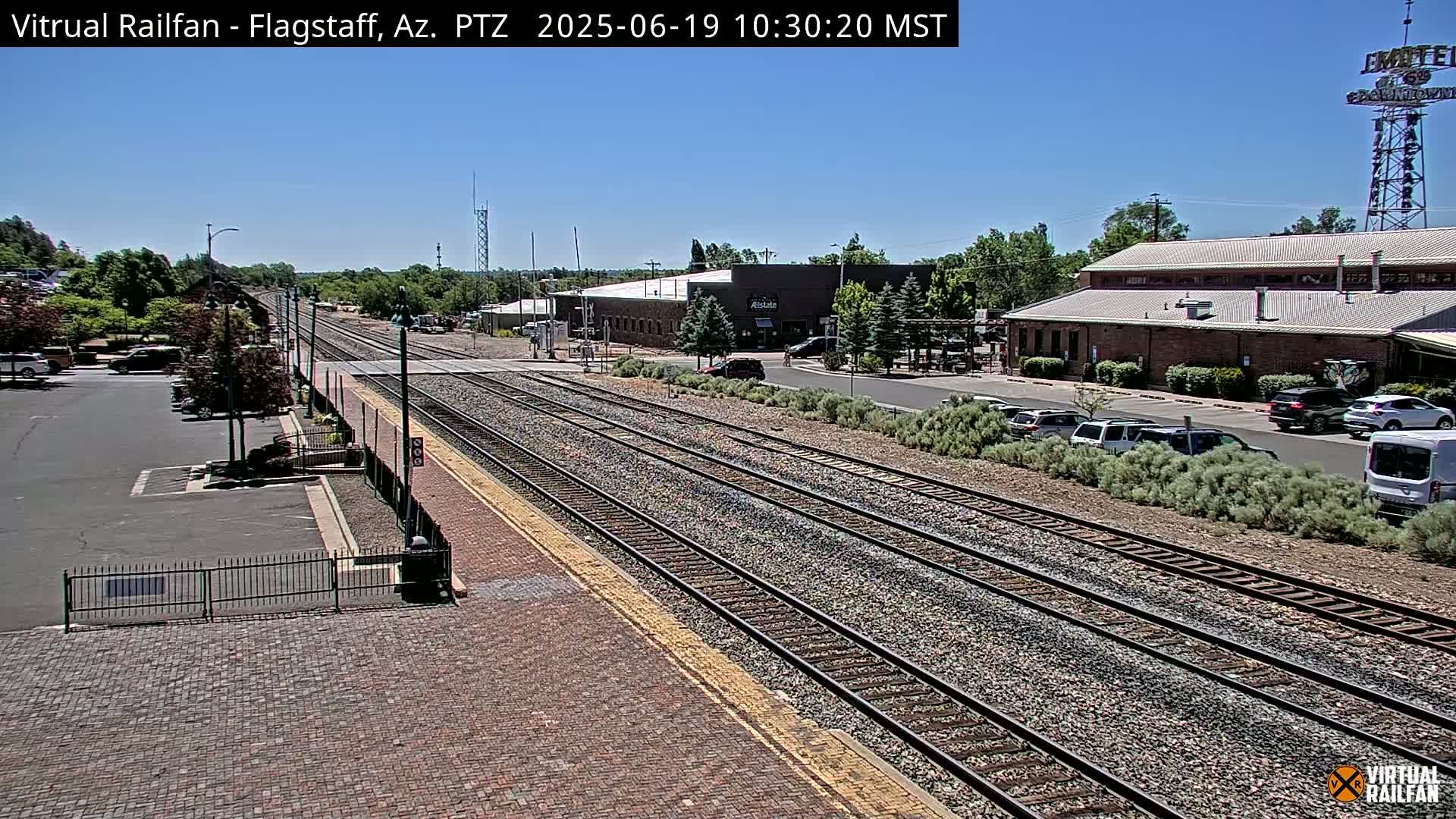 A train station with multiple tracks and parked cars under a clear, sunny sky.