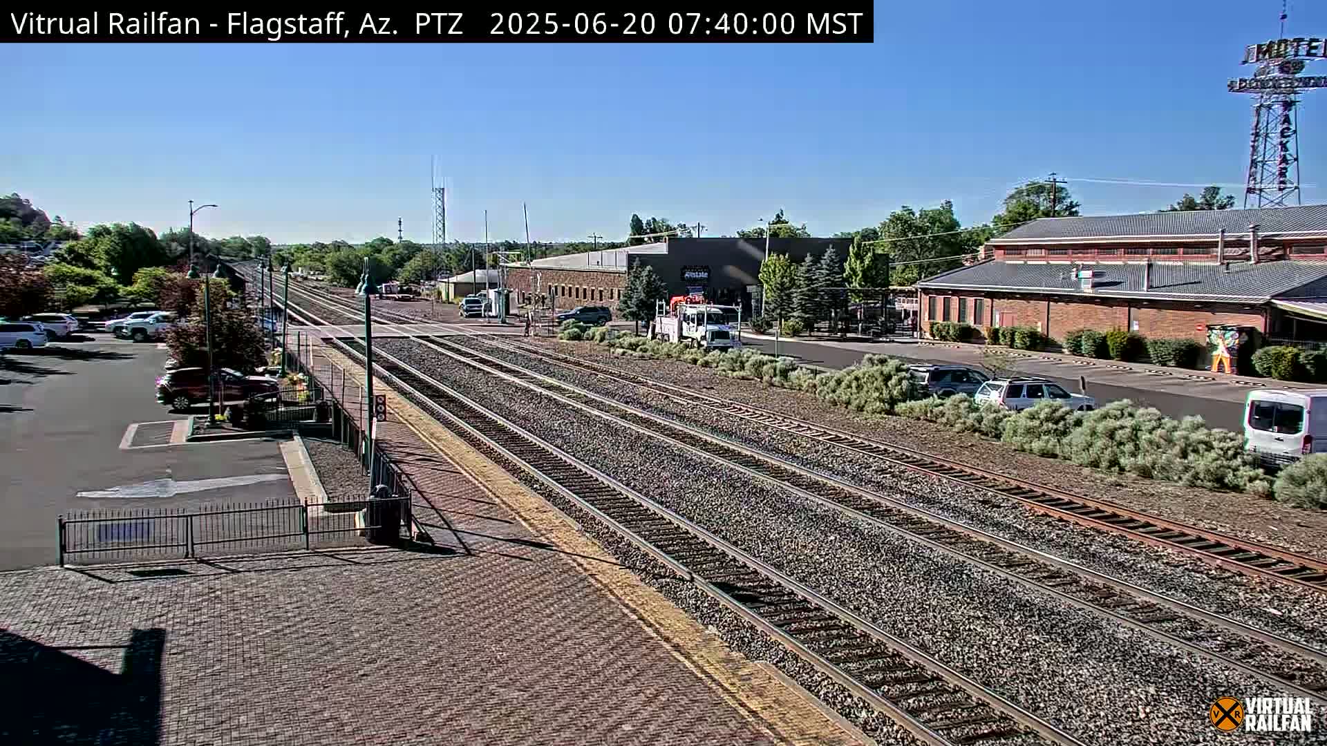 A sunny view of a train station with multiple train tracks, several parked cars, and buildings in the background.