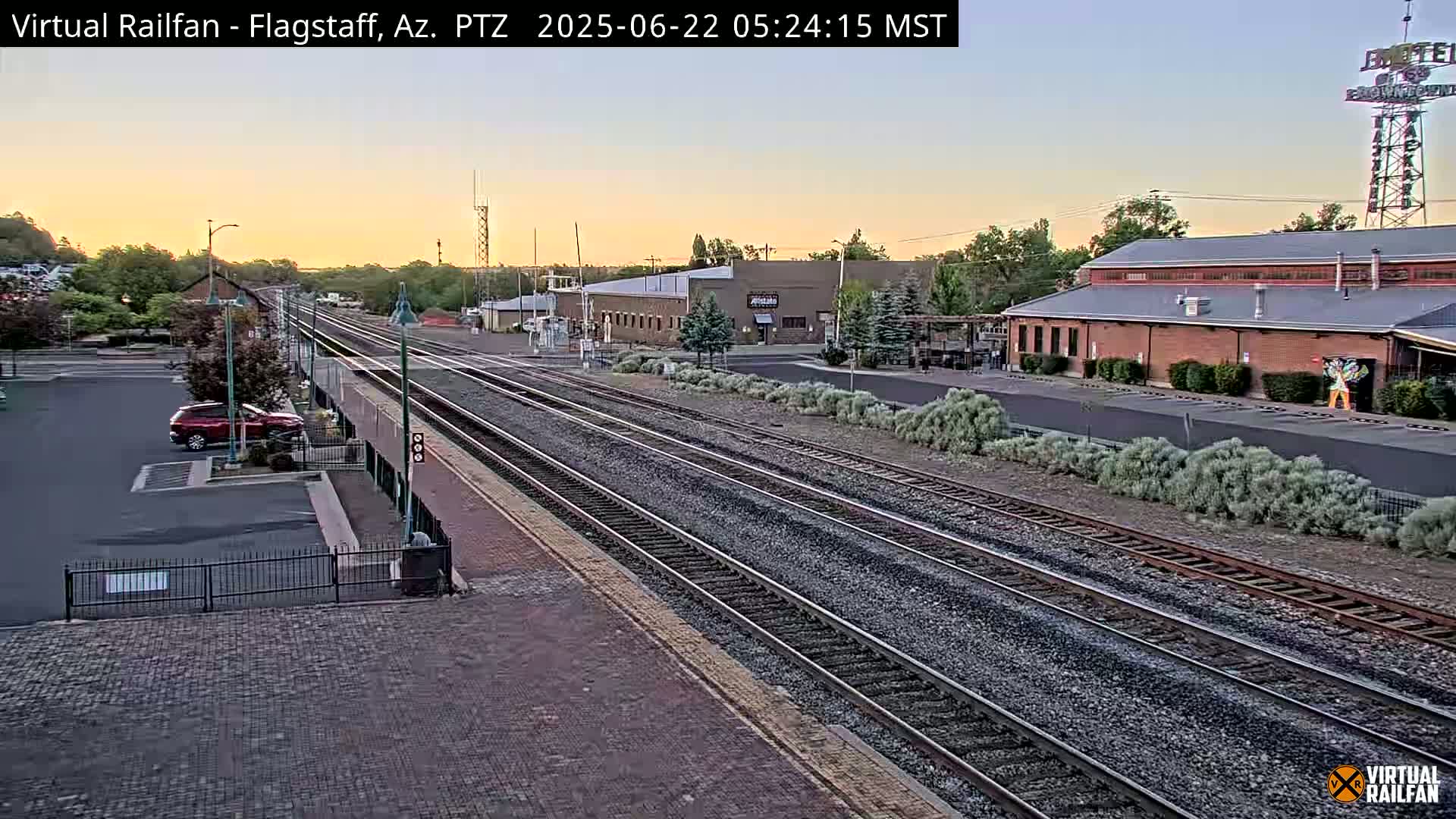 A train station at sunrise with multiple train tracks and a car parked in a nearby lot.